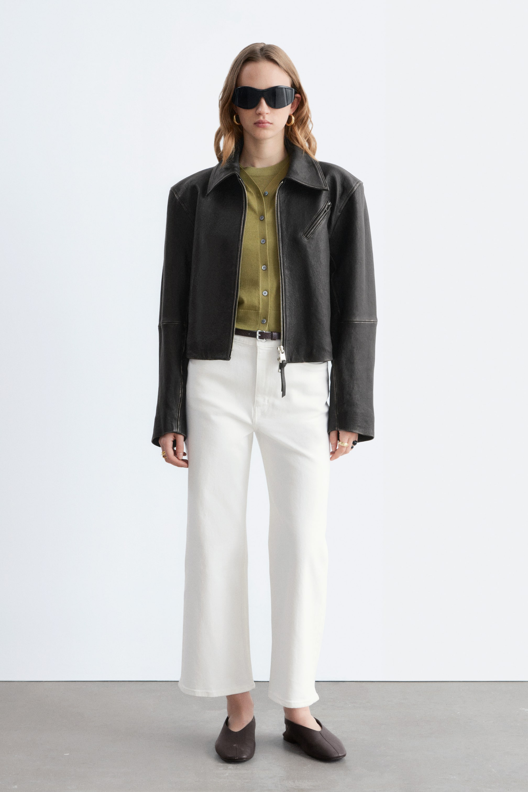 Zobrazit větší obrázek: Tmavě šedá kožená bunda s výrazným límcem, strukturovanými rameny a šikmou kapsou na zip na hrudi je navrstvená přes olivově zelený propínací svetr. Bílé široké kalhoty s oříznutou délkou jsou doplněny tmavě hnědým páskem a tmavě hnědé nazouvací baleríny dotvářejí celý outfit.
