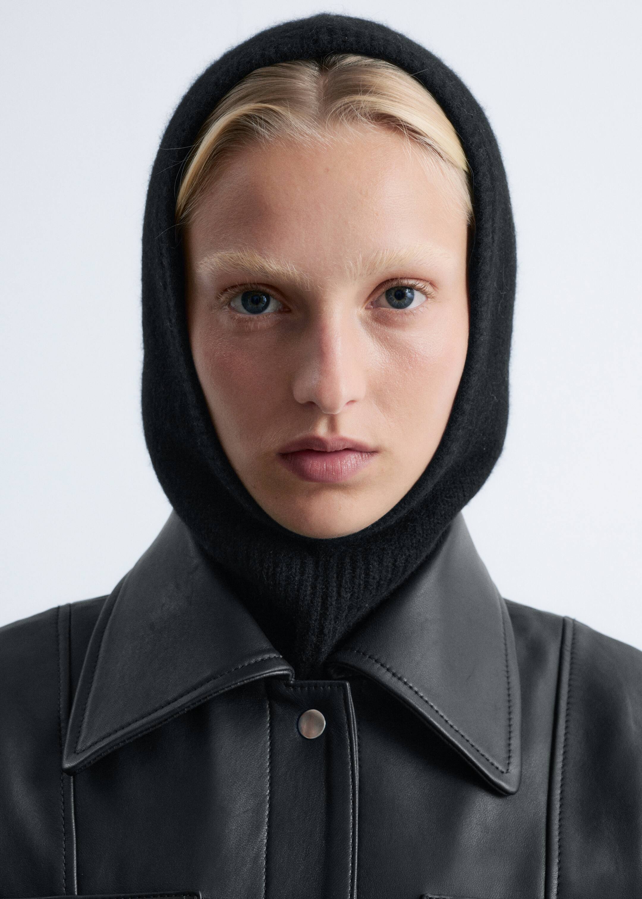 View larger image: A black knitted balaclava hood, featuring a fine texture and a ribbed section at the neck, is being worn by the model. It is framing the model's face and fitting snugly around the head and neck, layered under a black leather jacket.