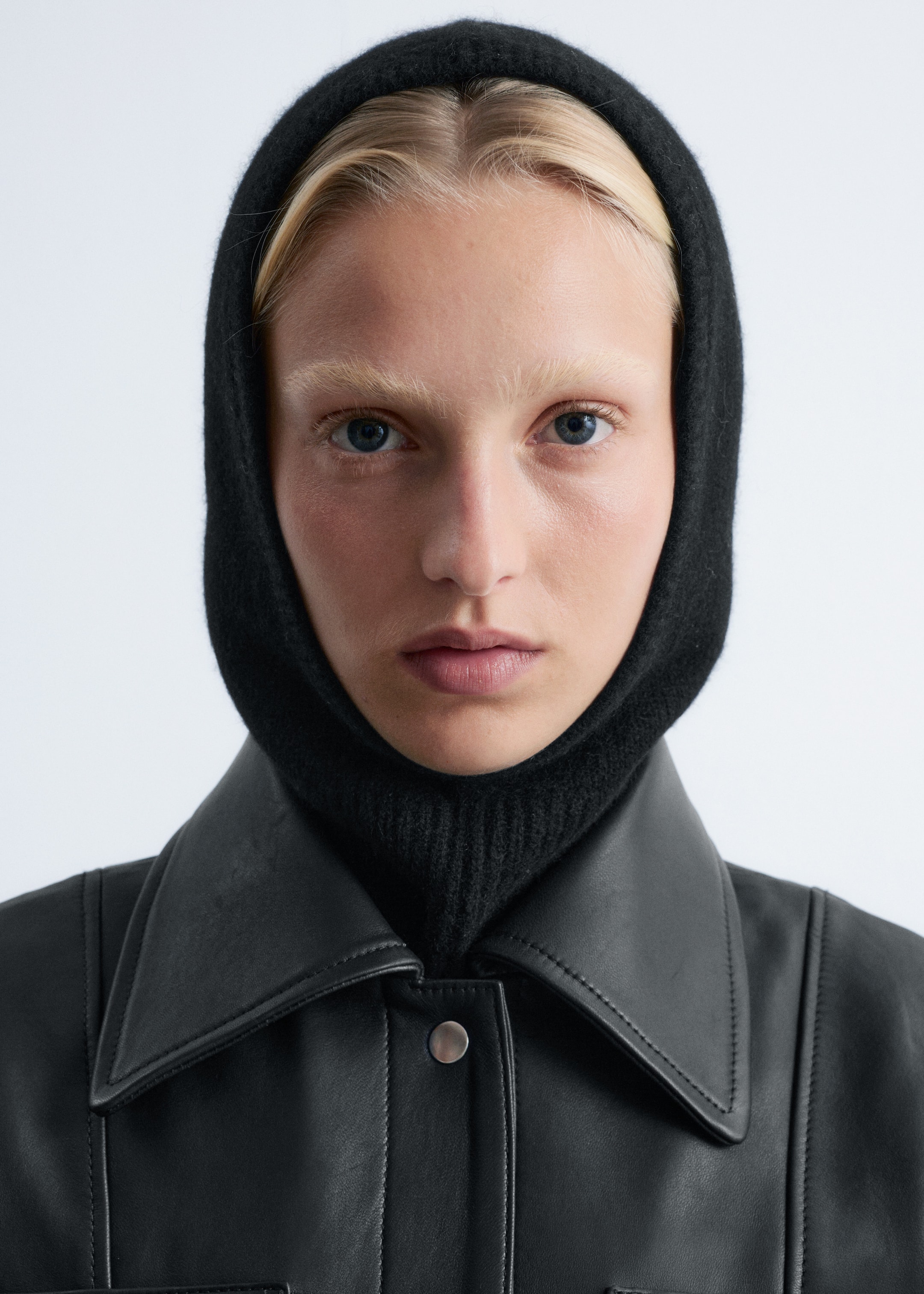 Vezi imaginea mai mare: O cagulă neagră tricotată, având o textură fină și o secțiune reiată la gât, este purtată de model. Aceasta încadrează fața modelului și se potrivește strâns în jurul capului și gâtului, fiind stratificată sub o jachetă de piele neagră.