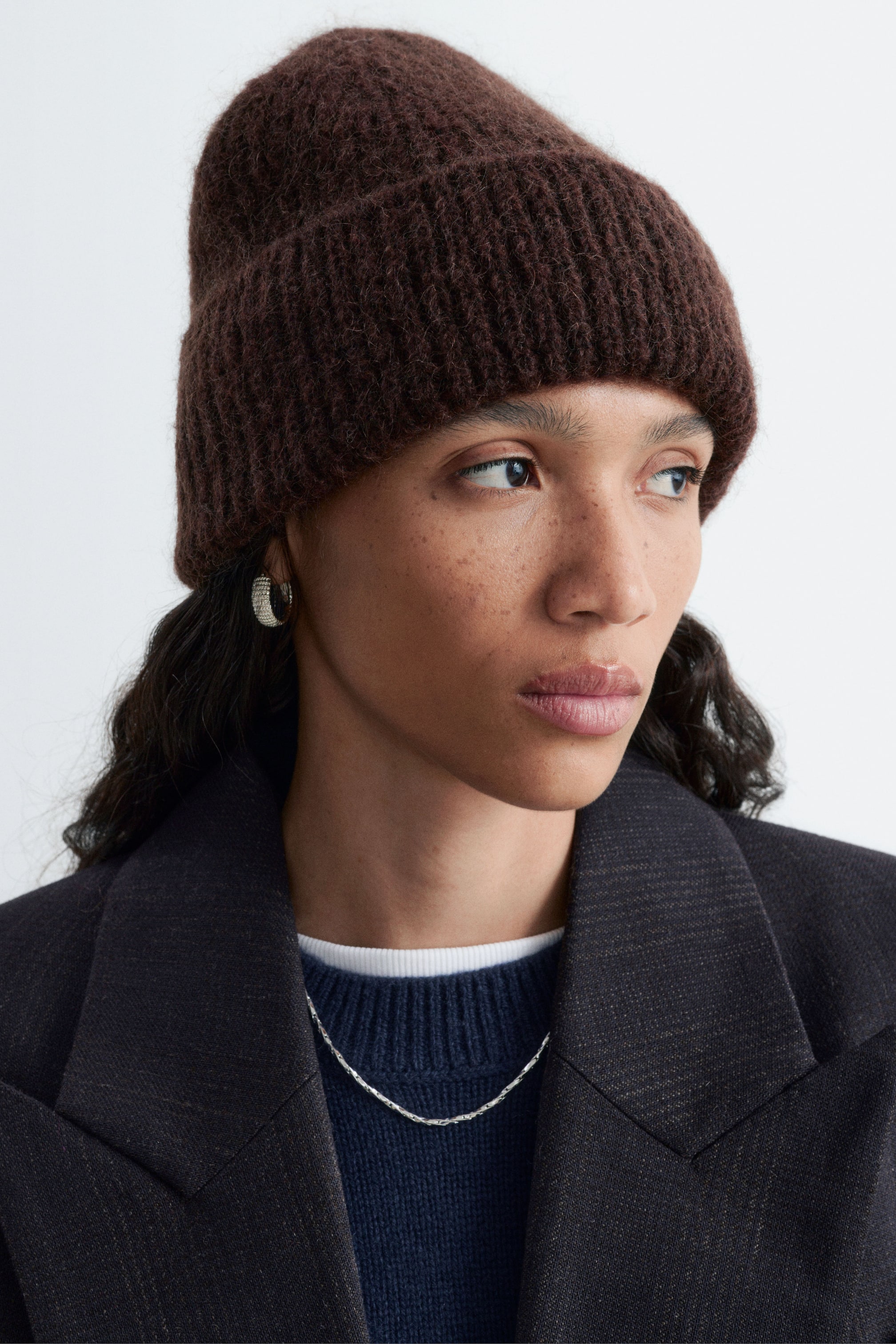 Agrandir l'image: Un bonnet côtelé texturé marron foncé est porté par le modèle, avec un large revers plié épais. Le bonnet est stylisé avec un blazer gris foncé superposé à un pull bleu à col rond.