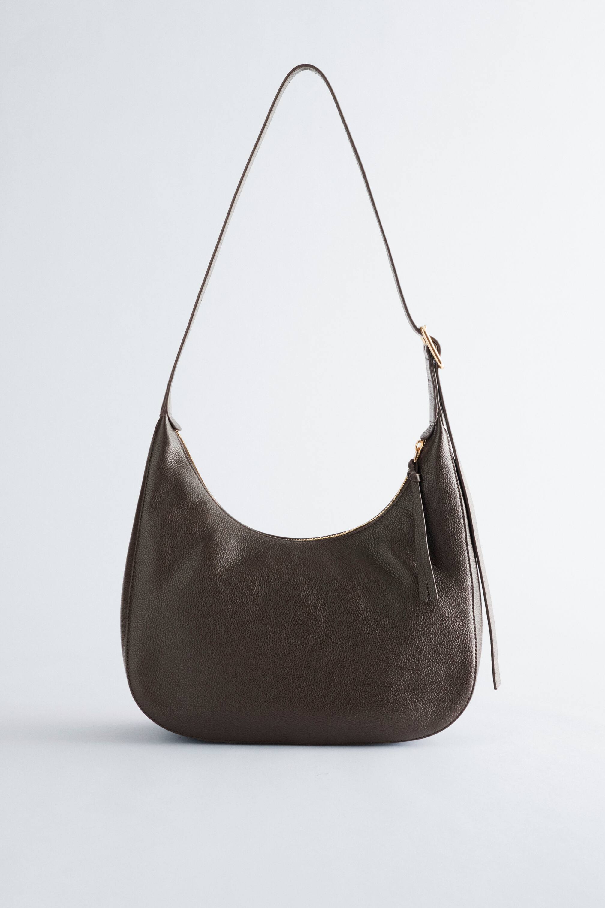 Nagyobb kép megtekintése: Egy sötétbarna, kavicsos bőr táska látható ELŐLRŐL, amely ívelt, laza sziluettet mutat. Hosszú, állítható vállpánttal rendelkezik, aranyszínű csattal, valamint felső cipzáras záródással és bőr húzófüllel.