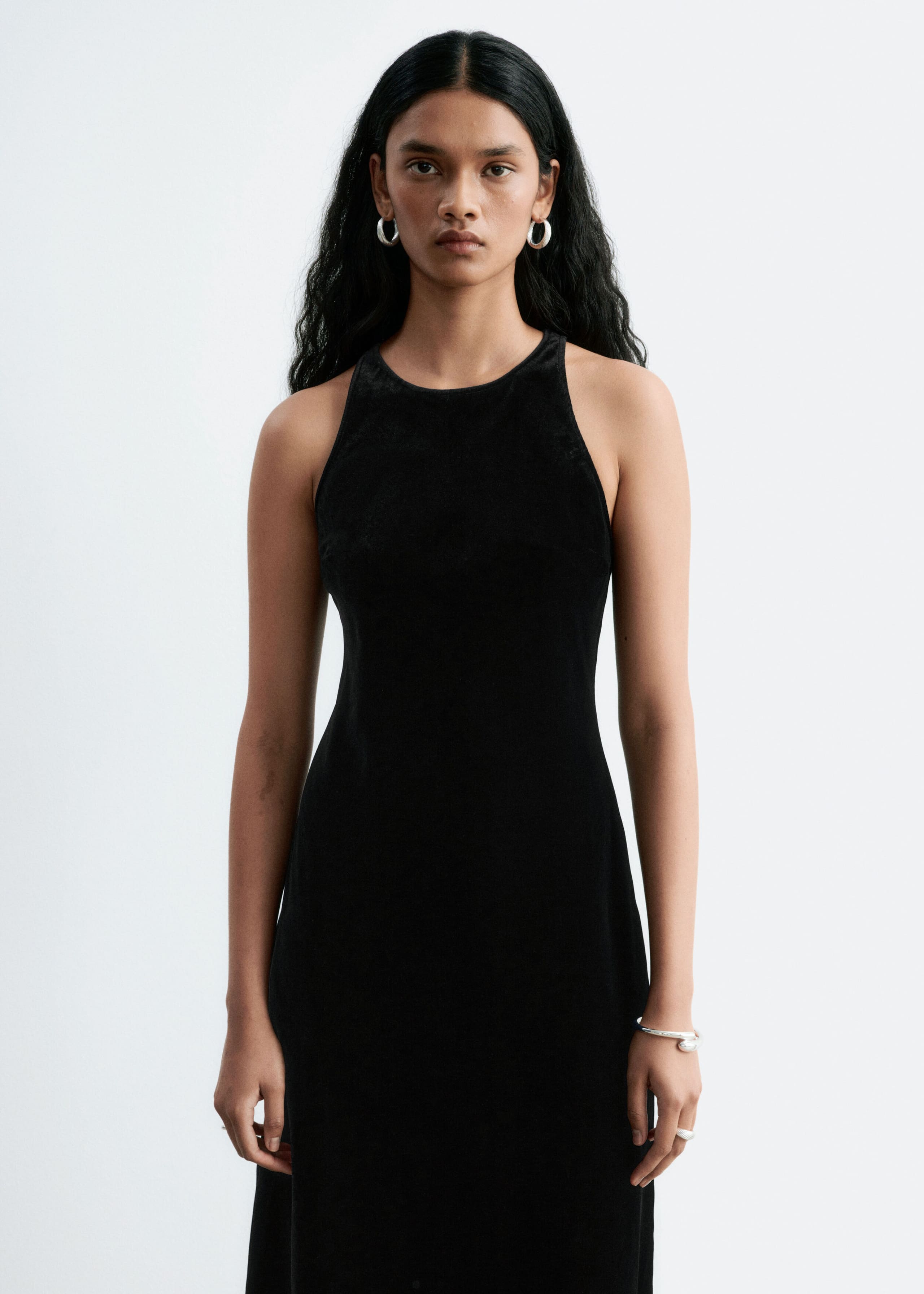 A black sleeveless dress with a high crew neckline is being worn by the model standing and facing forward. The dress is styled with silver hoop earrings, a silver bracelet, and a silver ring.