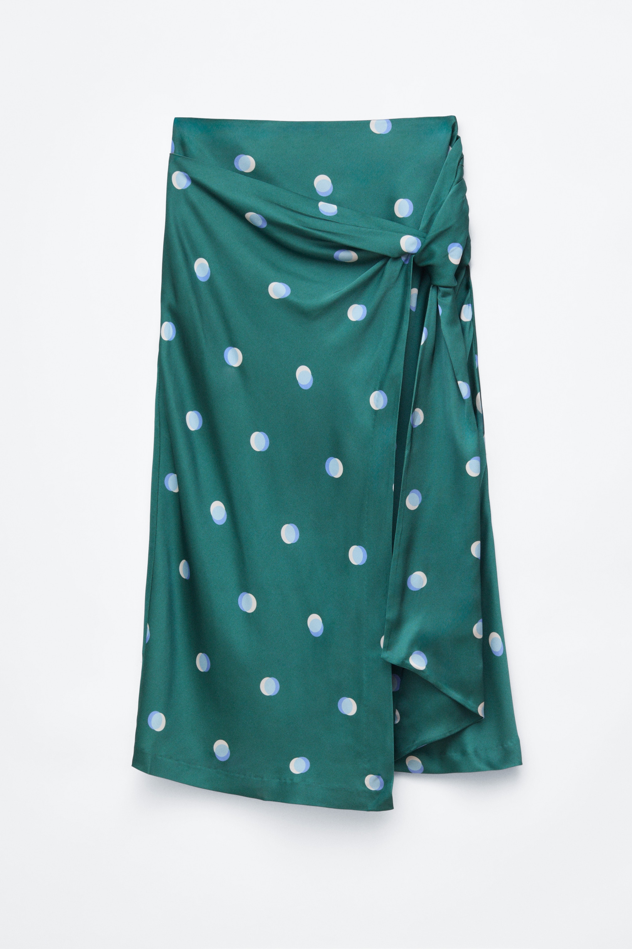 View larger image: A green midi skirt featuring a light blue and white polka dot pattern is shown from the front, highlighting a draped and knotted detail at the side waist. This draped construction creates a wrap-style front with an asymmetrical layered hem.
