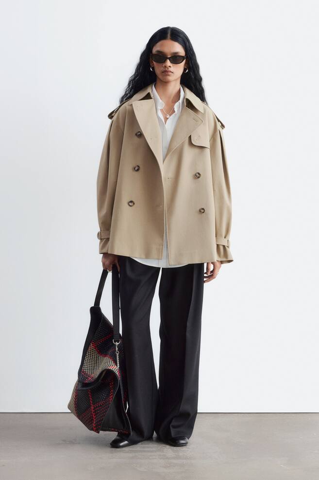 En beige kort trenchcoat bÃ¤rs av modellen, lager pÃ¥ lager Ã¶ver en vit skjorta med krage och svarta byxor med vida ben. Den dubbelknÃ¤ppta kappan har breda slag med notcherad kant, axelklaffar och remdetaljer vid manschetterna. - 1