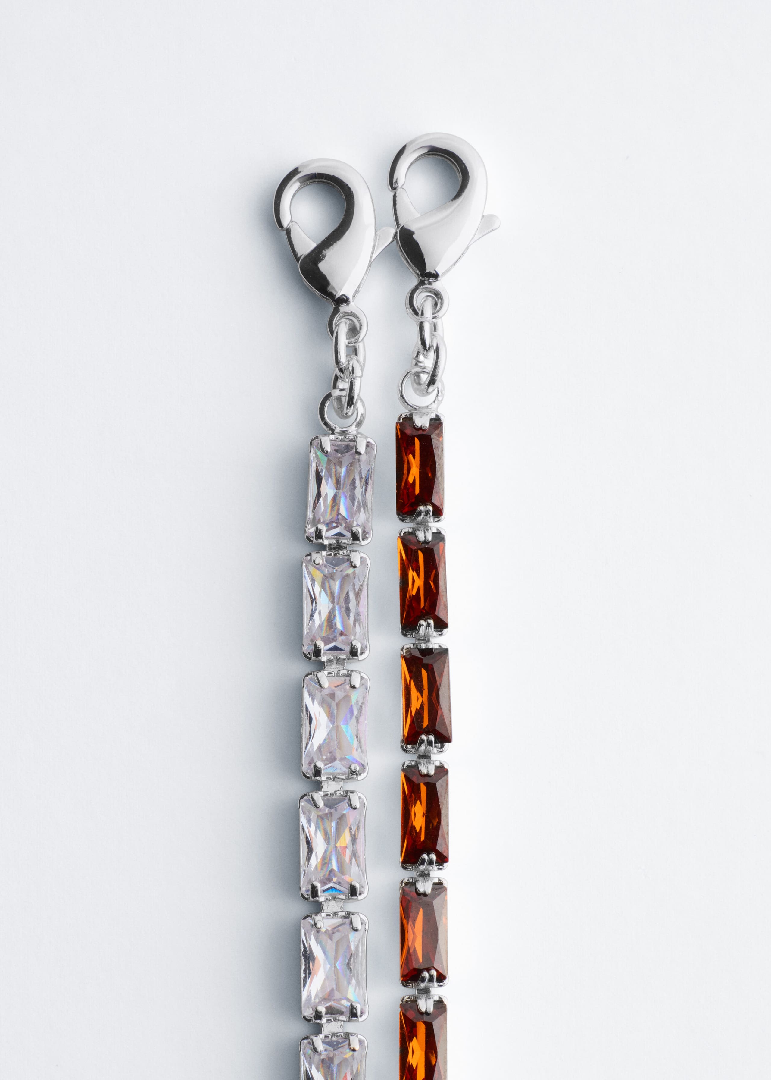 Two versions of the tennis chain necklace are hanging side by side, with one featuring clear, rectangular-cut cubic zirconia stones and the other featuring amber-colored, rectangular-cut stones. Both silver-toned necklaces are showing their lobster claw clasps at the top.