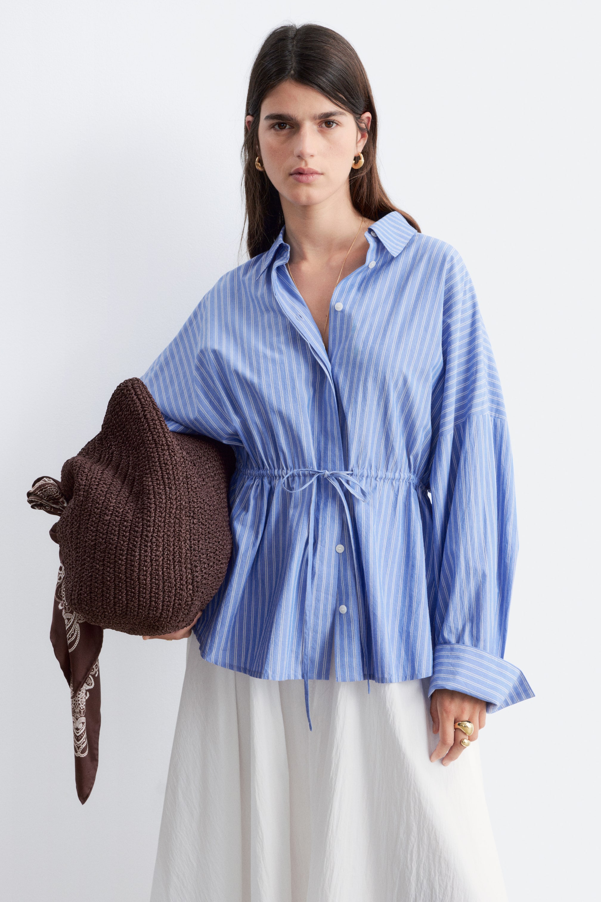 Zobrazit větší obrázek: Modro-bílá pruhovaná košile je vybavena špičatým límcem, zapínáním na knoflíky vpředu a objemnými rukávy s manžetami na knoflíky. Stahovací šňůrka v pase vytváří nabíraný peplum lem.