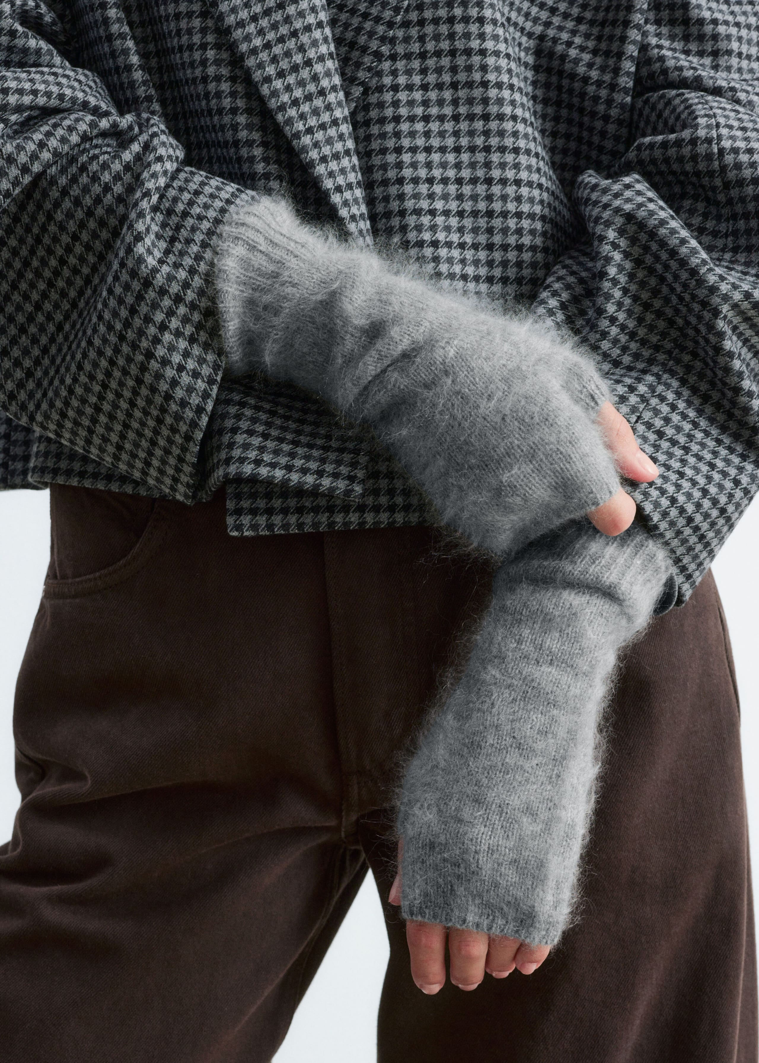 Grey fingerless gloves with a soft, brushed texture and ribbed cuffs are being worn by the model, who is clasping their hands together. The gloves are styled with a grey and black houndstooth jacket and brown trousers.