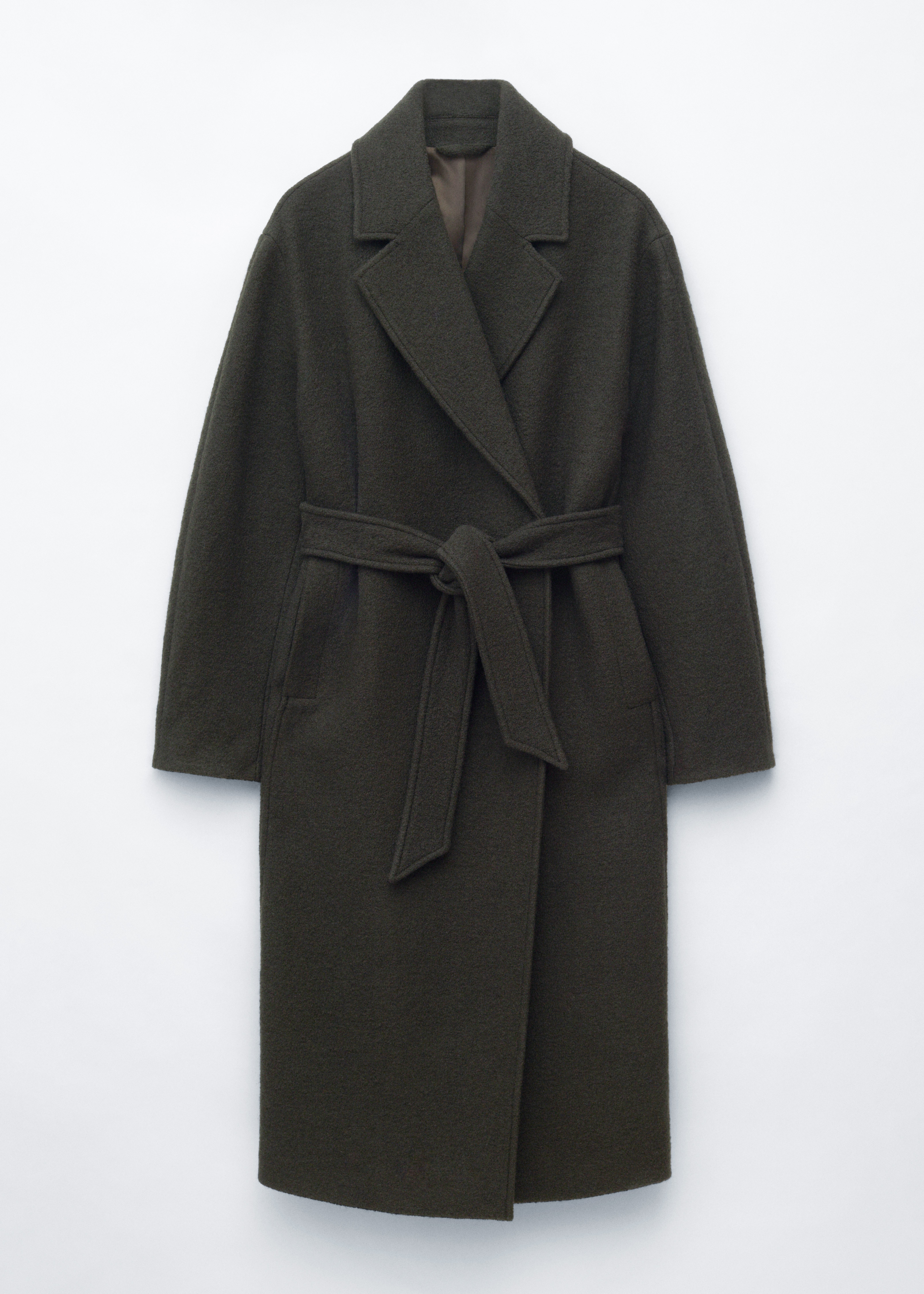 La modelo lleva un abrigo largo de doble botonadura en verde oscuro, con solapas de muesca y detalles de botones, sobre una camisa rosa claro y unos pantalones grises. La modelo lleva un bolso de hombro en topo y apoya una mano en el bolsillo lateral del abrigo.
