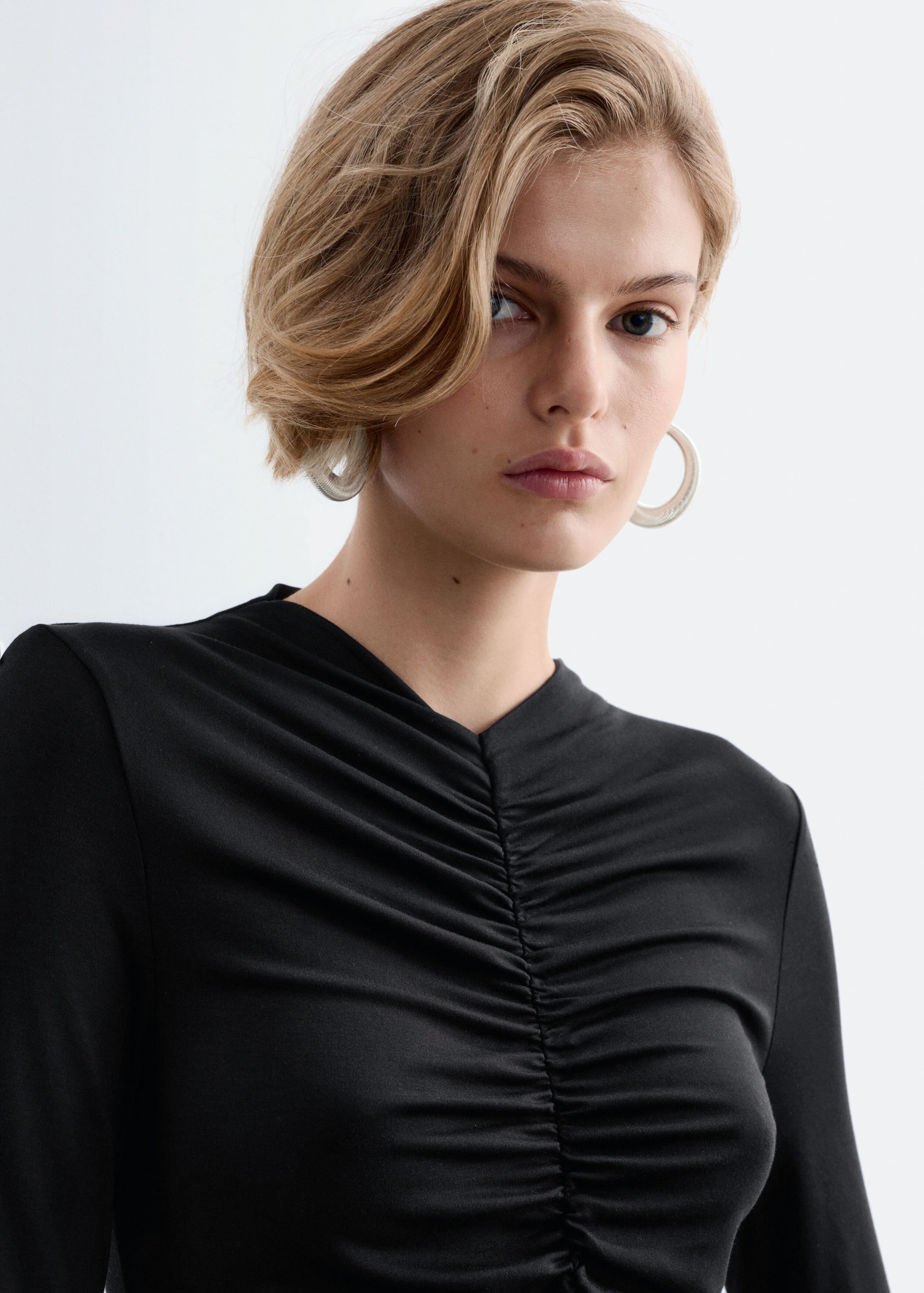 A black, close-fitting, long-sleeved top featuring prominent gathers along the center front seam is worn by the model. The jersey fabric is ruched across the chest, creating a textured drape.