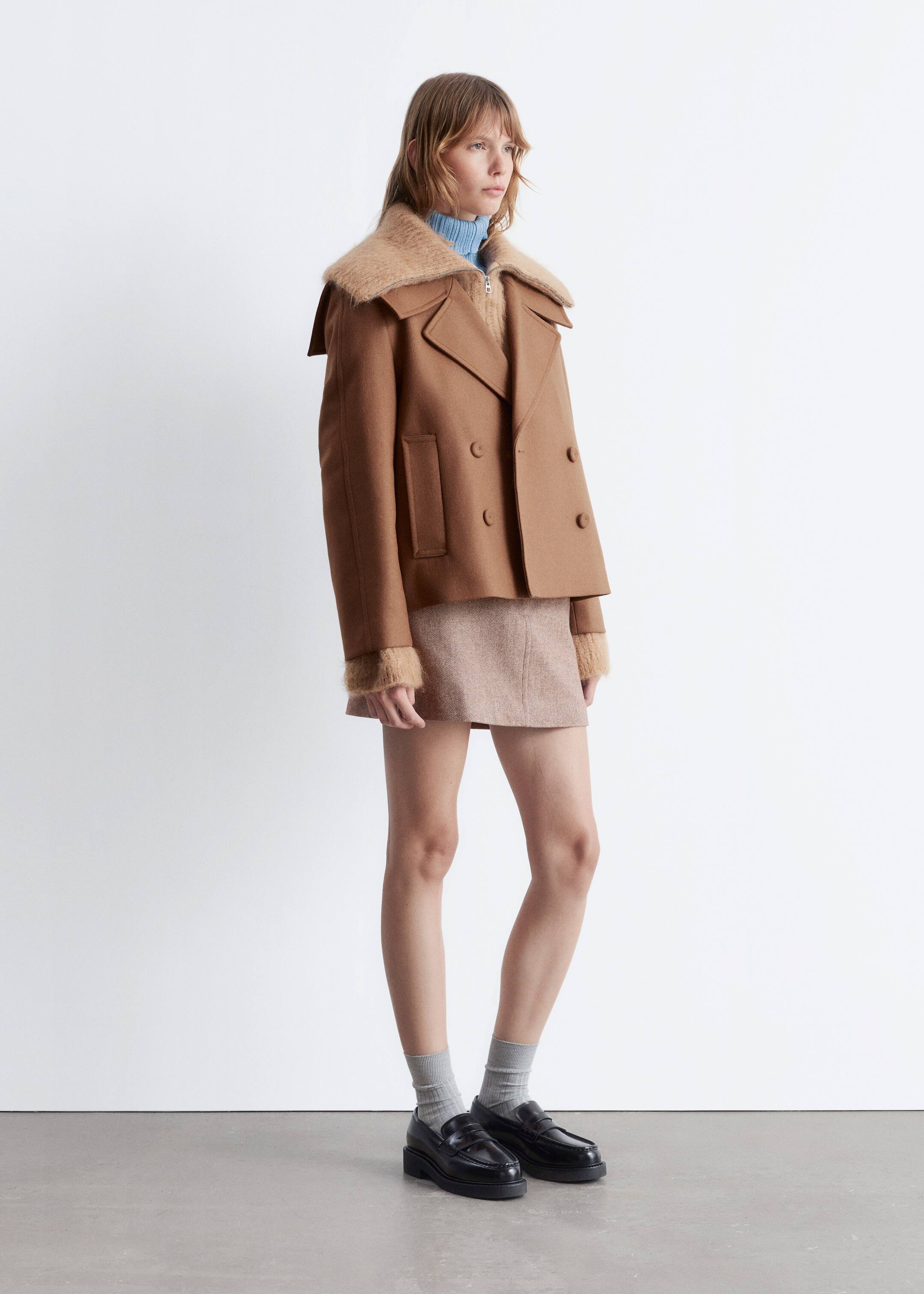 Agrandir l'image: Une veste de style caban marron avec un large col et des poignets en fourrure est portée par le mannequin. Elle présente un boutonnage croisé, des revers en pointe et deux poches passepoilées visibles, portée sur un pull à col roulé bleu clair et une jupe marron clair.