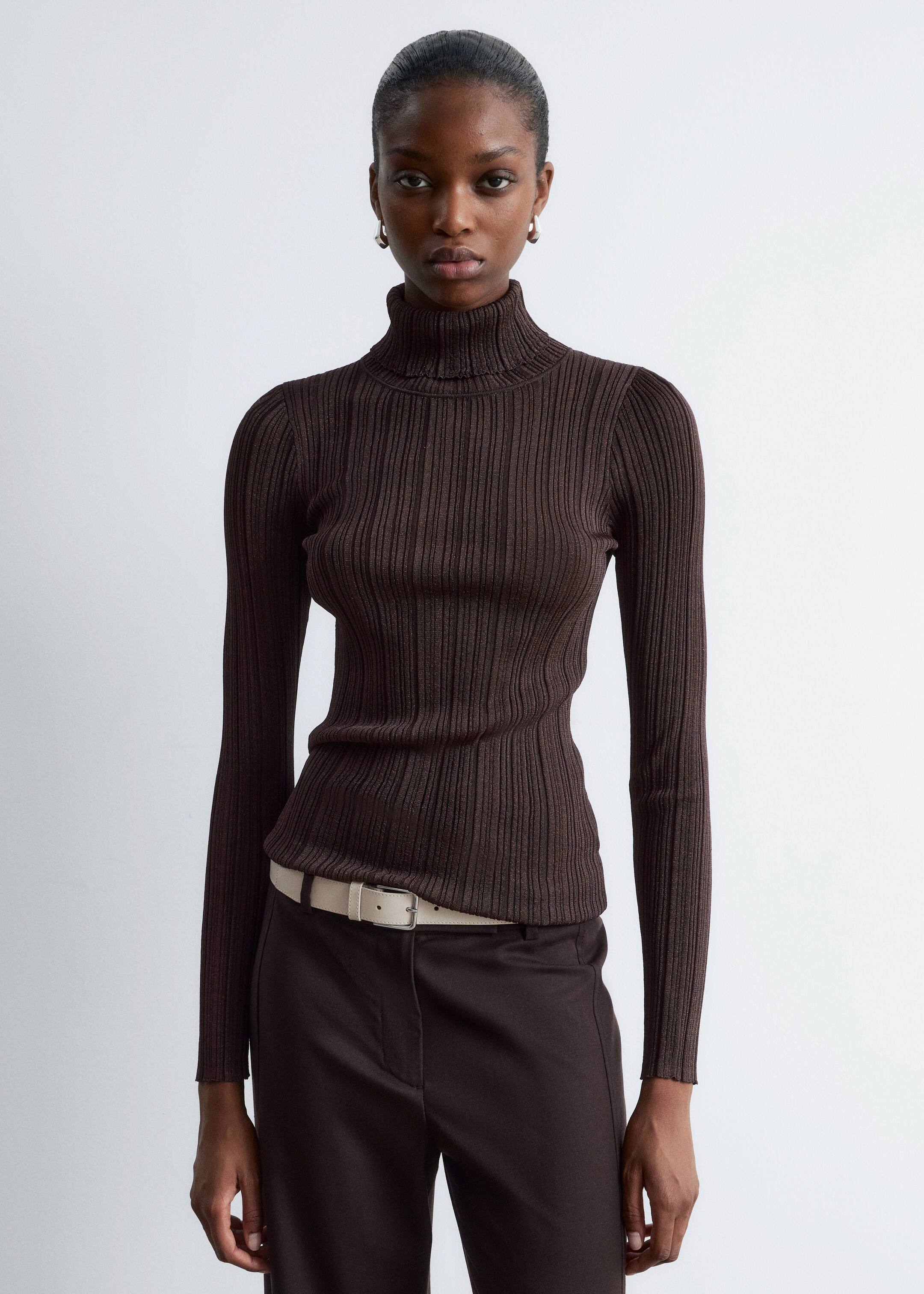 Ingrandisci l'immagine: La modella indossa un top a collo alto aderente, a coste fini, di colore marrone scuro, lavorato a maglia, con maniche lunghe e un sottile riflesso metallico, infilato in pantaloni marrone scuro abbinati a una cintura di colore chiaro.