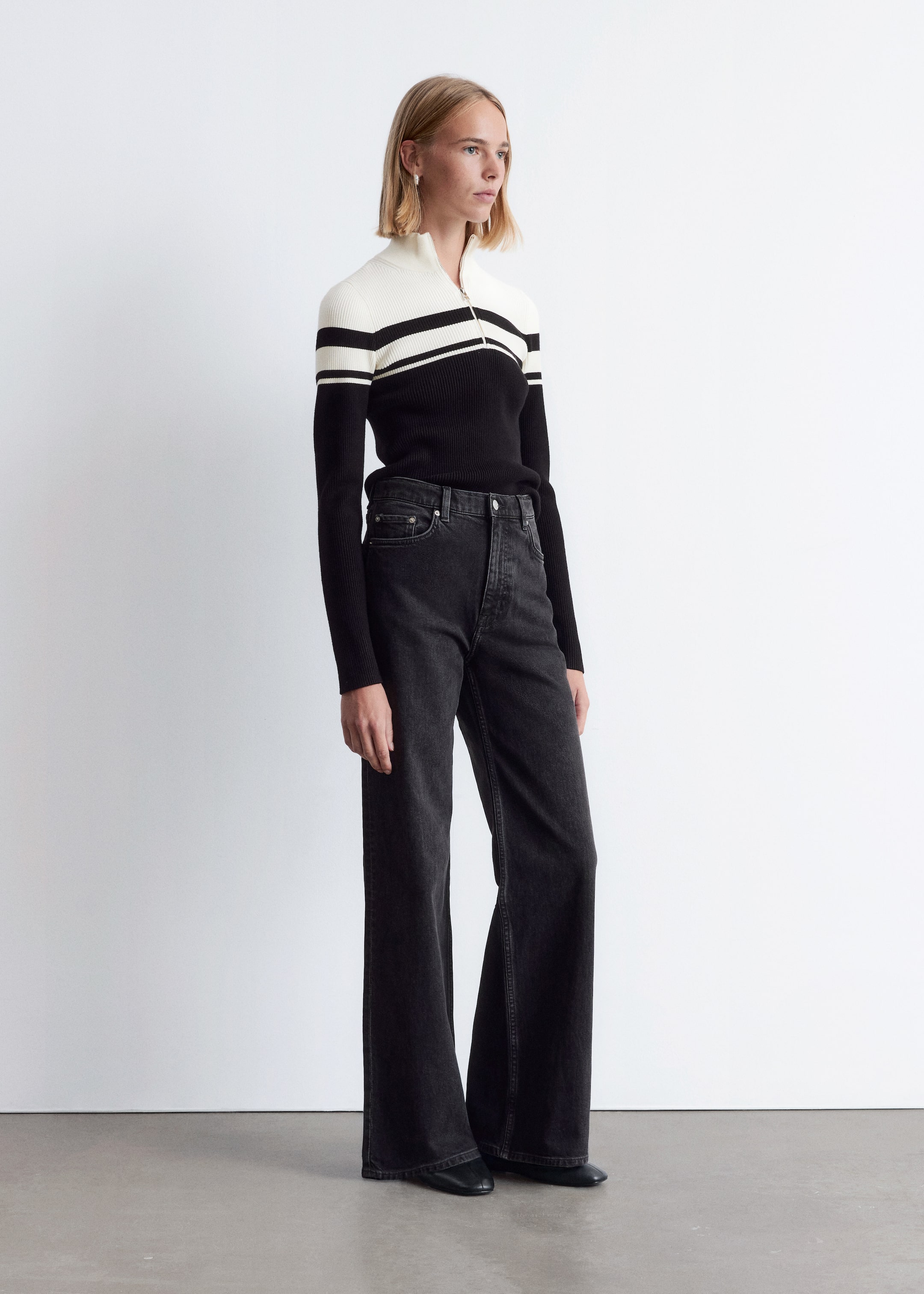 View larger image: A ribbed knit sweater, featuring a half-zip neckline, black lower body and long sleeves, and an off-white upper section with two horizontal black stripes, is being worn tucked into high-waisted, washed black wide-leg jeans. The jeans are presenting a relaxed silhouette and are covering black shoes.