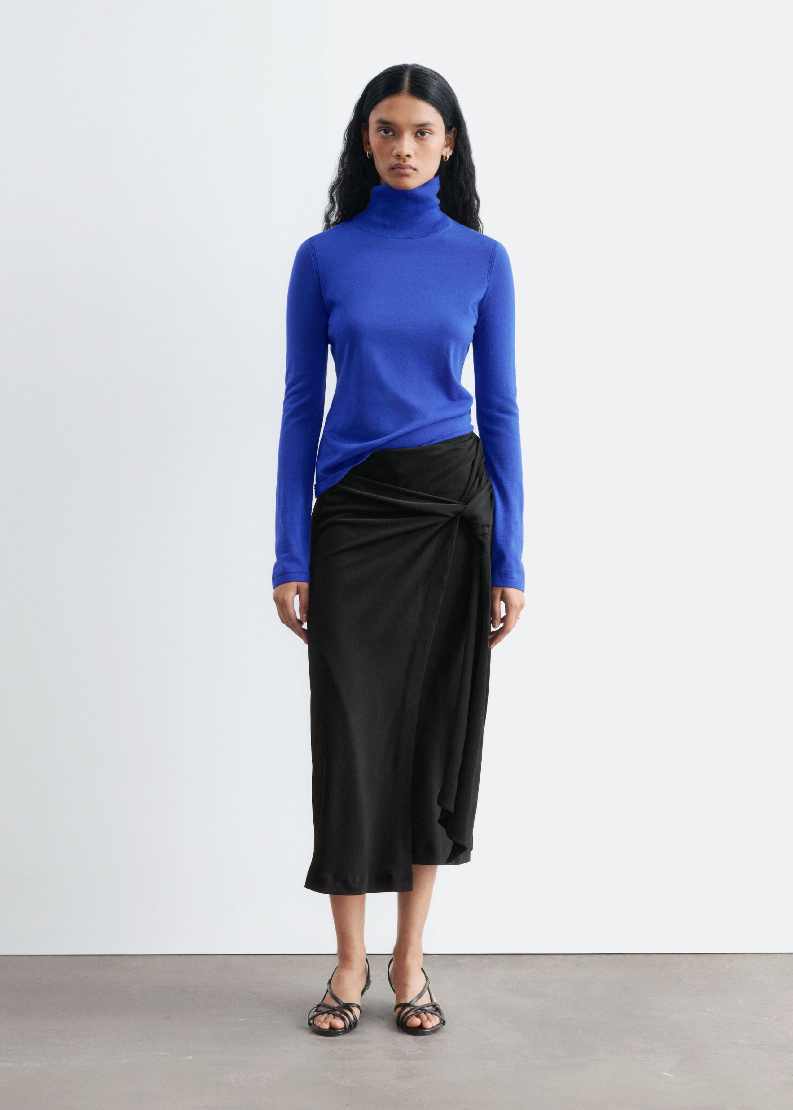 Het model draagt een koningsblauwe merinowollen coltrui met lange mouwen en een hoge kraag. De slim-fit top is gestyled met een gedrapeerde zwarte midi-rok.