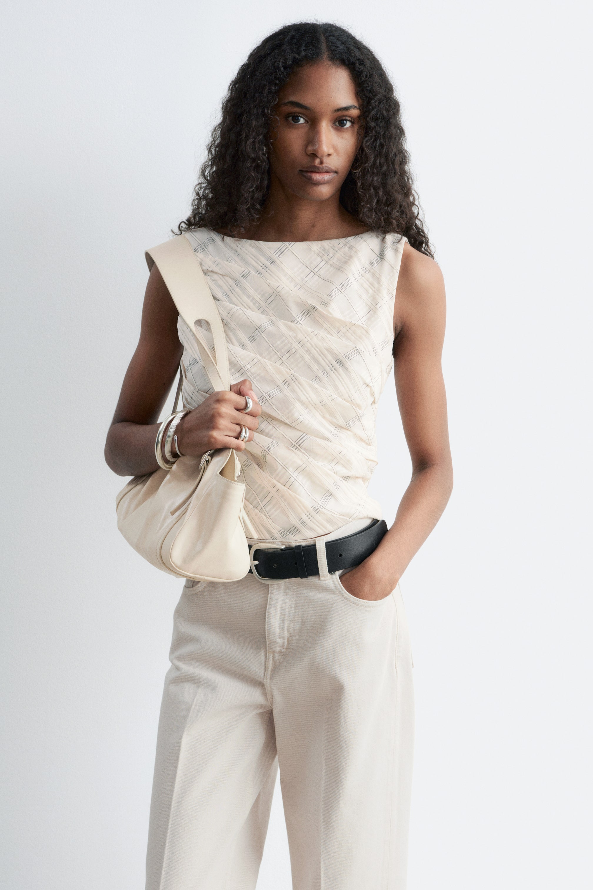 Agrandir l'image: Le haut sans manches présente un col bateau et un tissu couleur crème avec un motif à carreaux subtil. Il affiche des fronces et des drapés en diagonale sur le devant, avec son ourlet soigneusement rentré dans un pantalon et ceinturé par une ceinture noire.