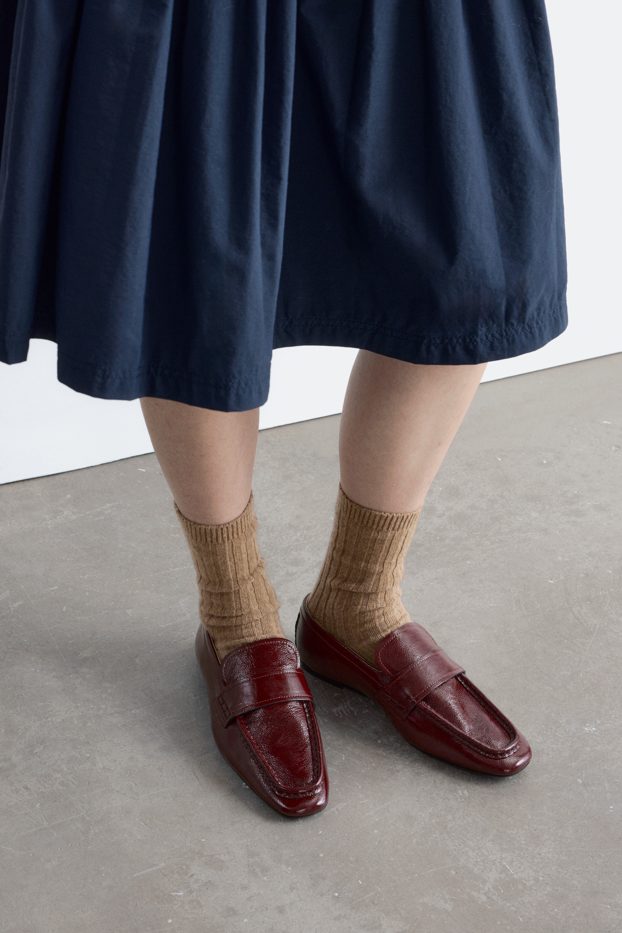 Ingrandisci l'immagine: Mocassini in pelle verniciata rosso scuro, caratterizzati da una punta quadrata e una fascetta da penny loafer, indossati con un'inclinazione leggermente in avanti e verso l'esterno, abbinati a calze beige a coste e un orlo di abito blu navy visibile sopra.