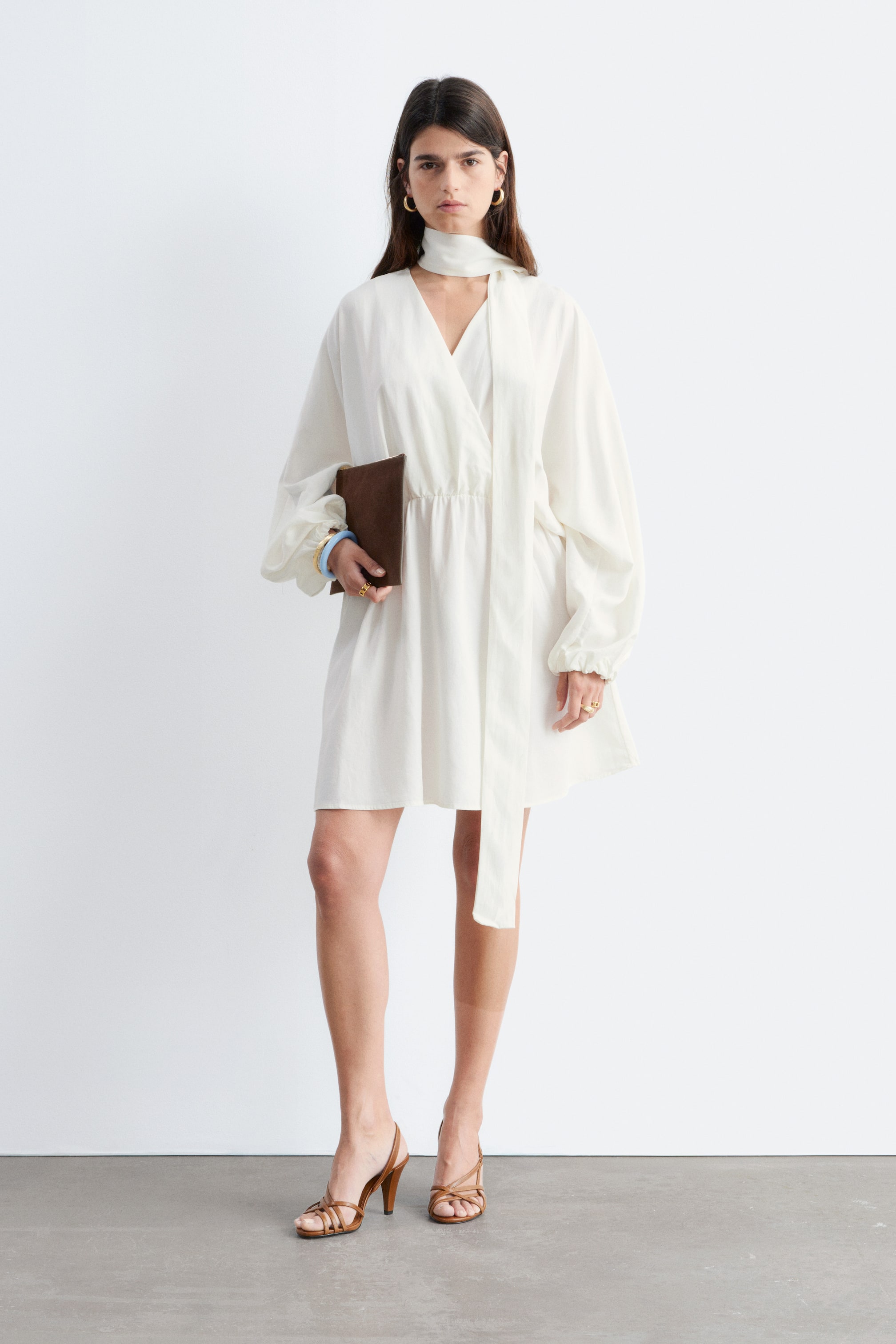 Zobrazit větší obrázek: Bílé mini šaty s překříženým výstřihem do V s dlouhou kravatou, objemnými dlouhými rukávy s nařasenými manžetami a elastickým pasem. Sukně je splývavá a dosahuje krátké délky.