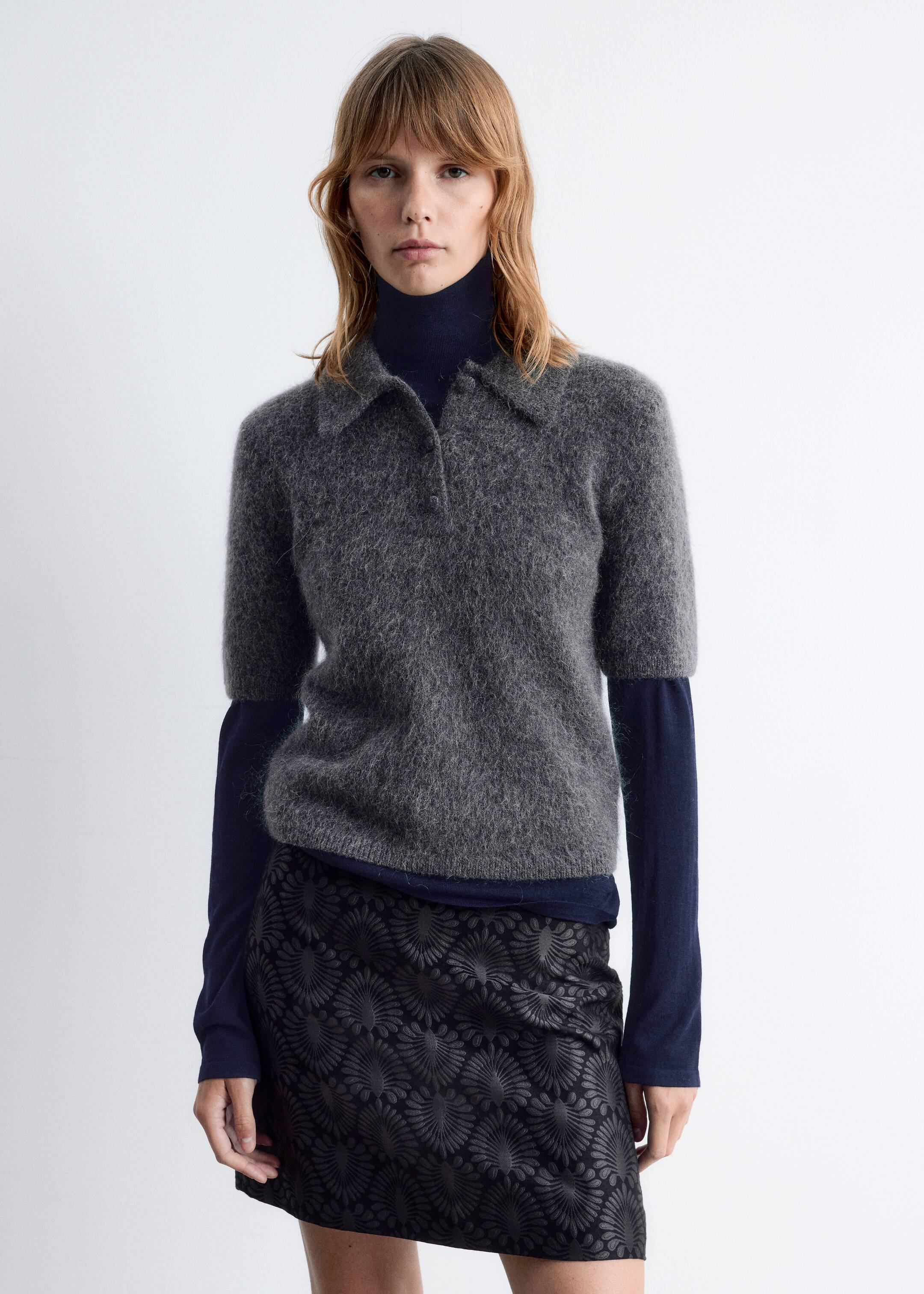 Größeres Bild ansehen: Ein graues, flauschiges, kurzärmeliges Polo-Top mit einem spitzen Kragen und einer Zwei-Knopf-Leiste wird aus einer frontalen Perspektive präsentiert. Es wird über einem dunkelblauen langärmeligen Rollkragenpullover getragen und mit einem schwarzen Minirock mit geprägtem Fächermuster kombiniert.
