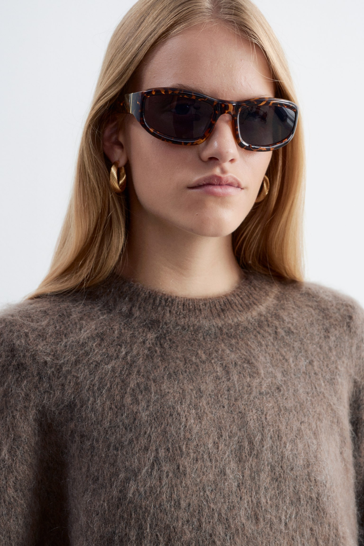 Een zachte, bruine trui van mohairmix met een geribbelde ronde hals wordt gedragen door het model, dat is gestyled met een zonnebril met schildpadmotief en gouden oorringen. - 1