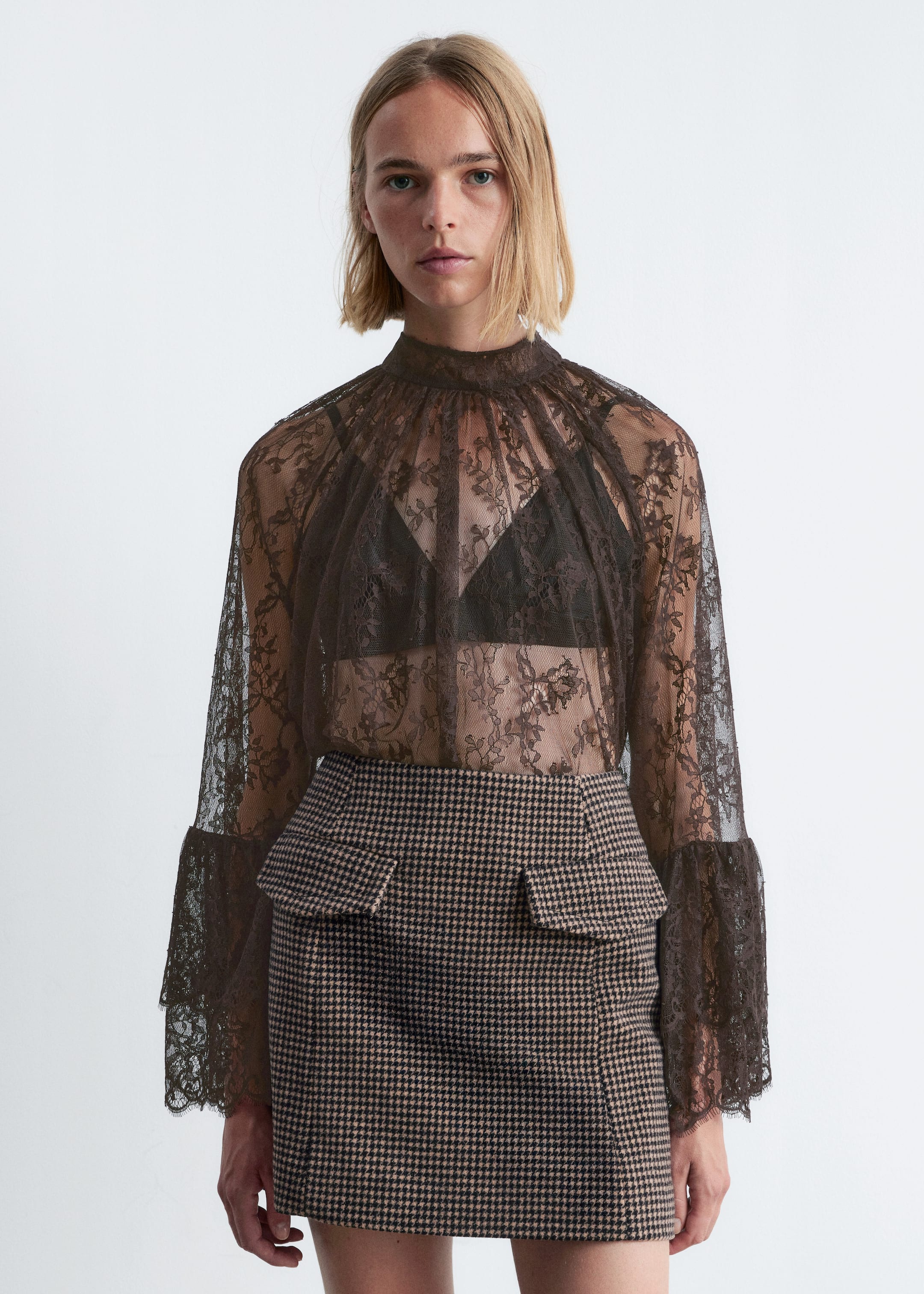View larger image: A dark brown sheer lace top, featuring a high mock neck and long sleeves with flared, ruffled lace cuffs, is being worn. A dark bra is visible underneath the top, which is being paired with a beige and black houndstooth mini skirt that has two angled front flap pockets.