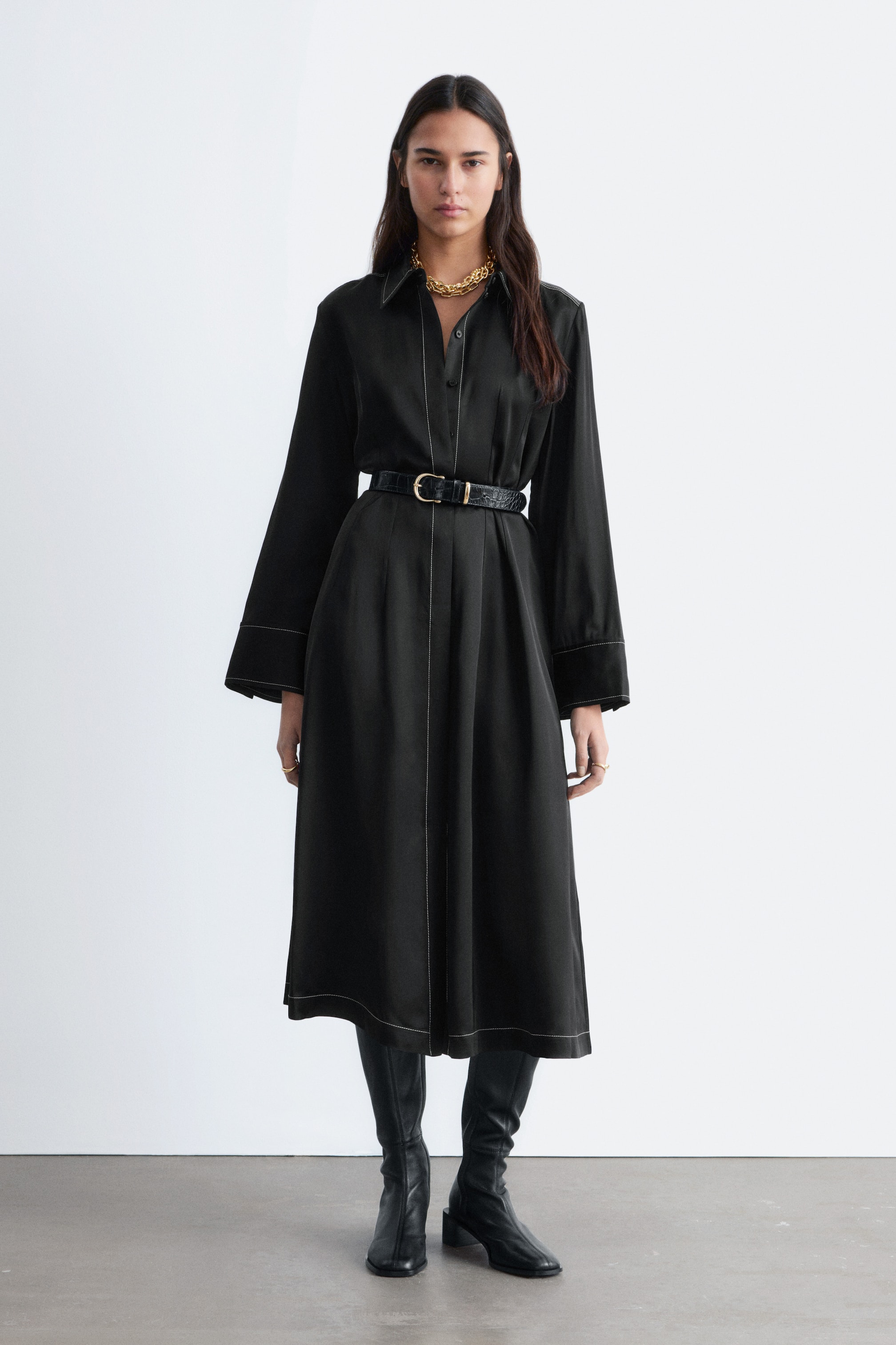 Agrandir l'image: Une robe-chemise midi noire est présentée, avec des manches longues et un col pointu. Elle est cintrée à la taille par une ceinture texturée noire, et des surpiqûres blanches contrastantes mettent en valeur ses coutures, poignets et ourlet, créant une silhouette en ligne A.