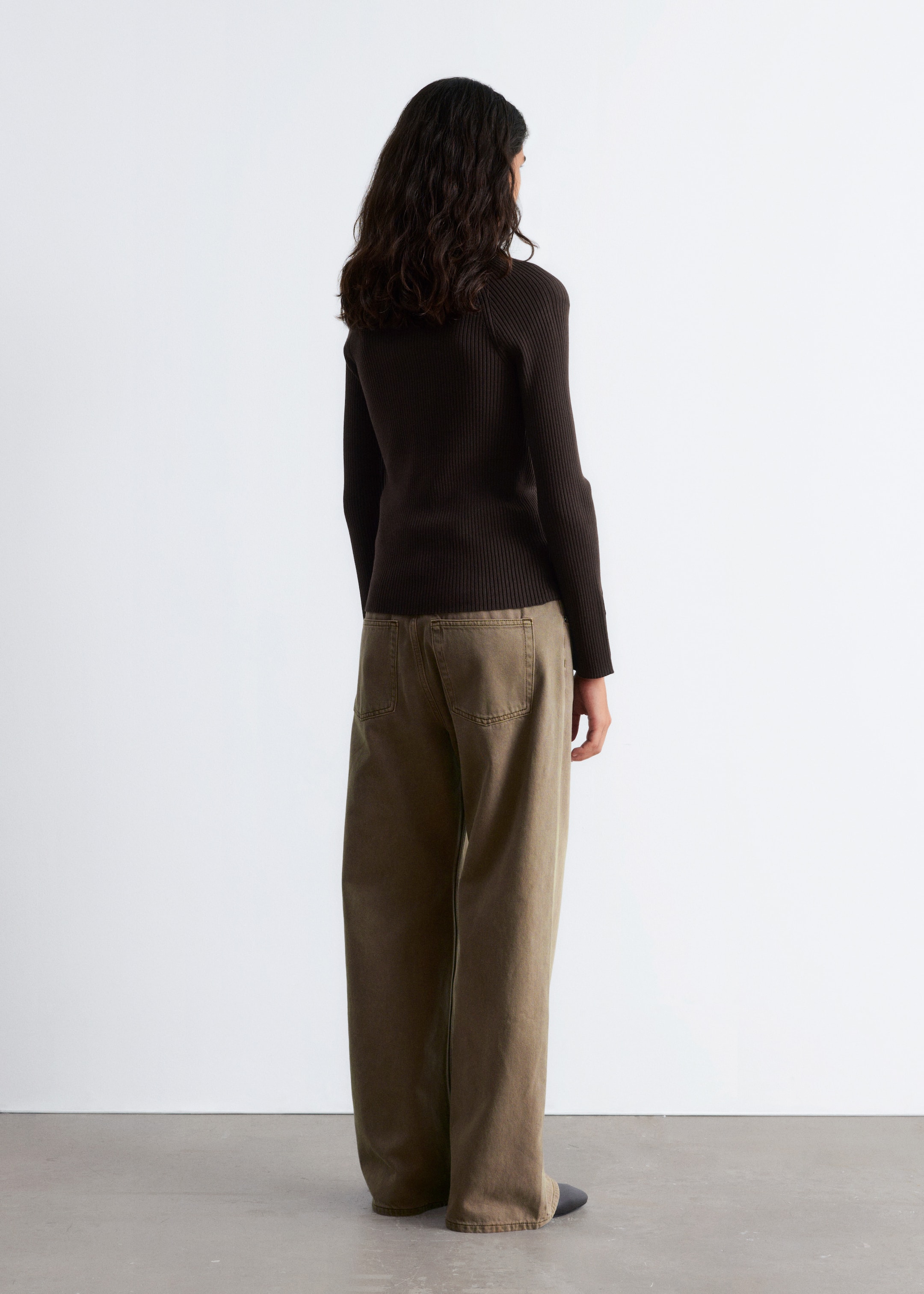 Grotere afbeelding bekijken: Een donkerbruin, geribbeld gebreid topje wordt gedragen door het model, getoond van de achterkant. Het topje heeft lange mouwen en een slank silhouet, en is gestyled met olijfkleurige wijde broek.