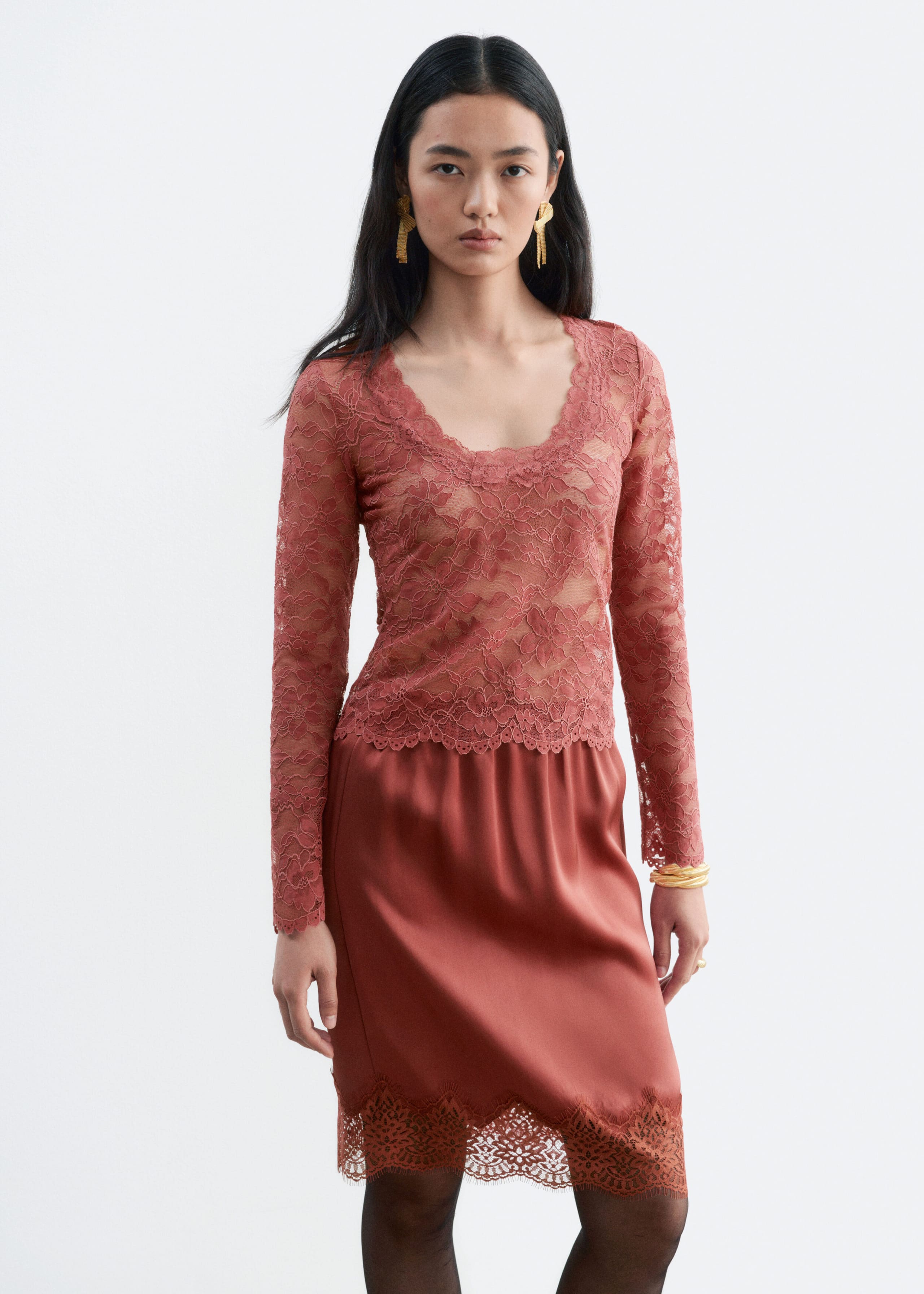 The model is wearing a long-sleeved, rose-coloured lace top with a scoop neckline, paired with a matching satin skirt featuring a lace-trimmed hem. The slim-fit top has scalloped edges at the cuffs and hem, while the skirt falls to the knee.