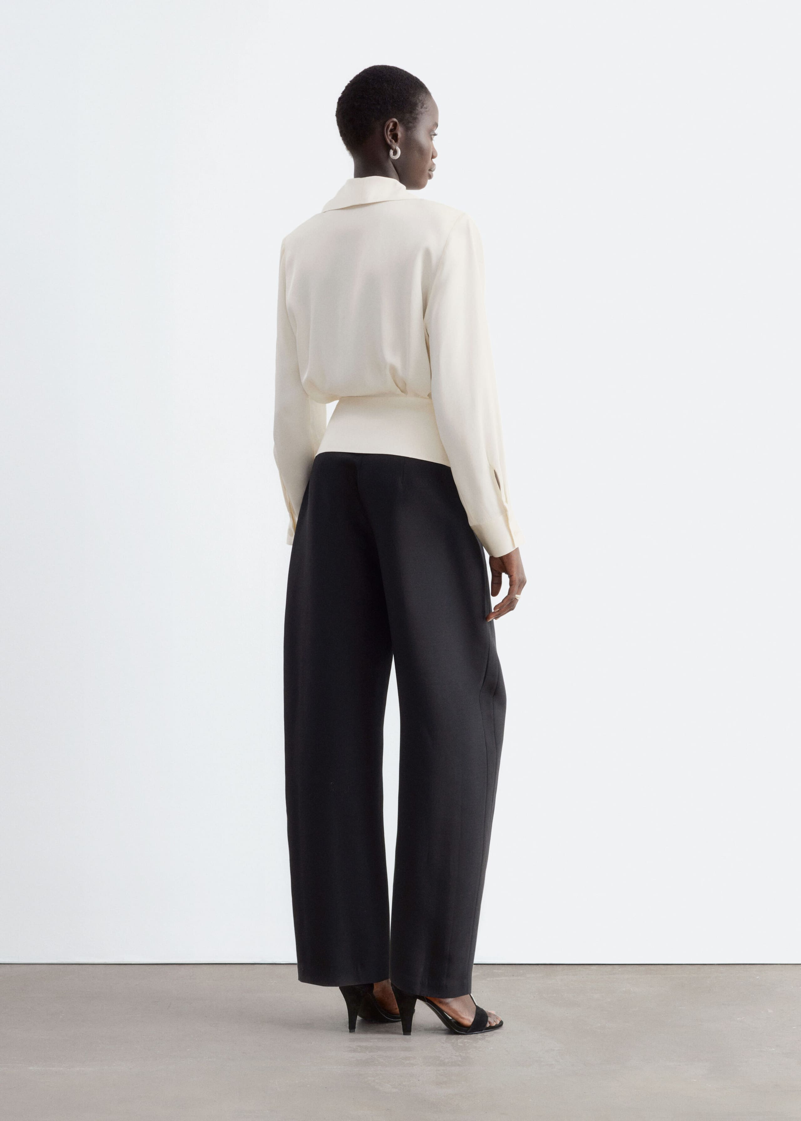La modella indossa una blusa avvolgente color crema a maniche lunghe, che mette in risalto i dettagli sul retro, tra cui un colletto a scialle e un punto vita arricciato. La blusa è abbinata a pantaloni neri a gamba larga e sandali neri con tacco.