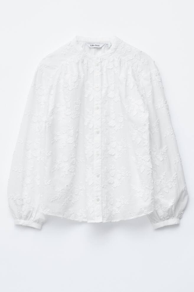 Eine weiße voluminöse Bluse wird VORNE präsentiert, mit einem Stehkragen, einer Knopfleiste vorne und einem durchgehenden Blumenapplikationsmuster. Sie hat lange Ärmel, die in gerafften Manschetten enden, und einen abgerundeten Saum. - 1