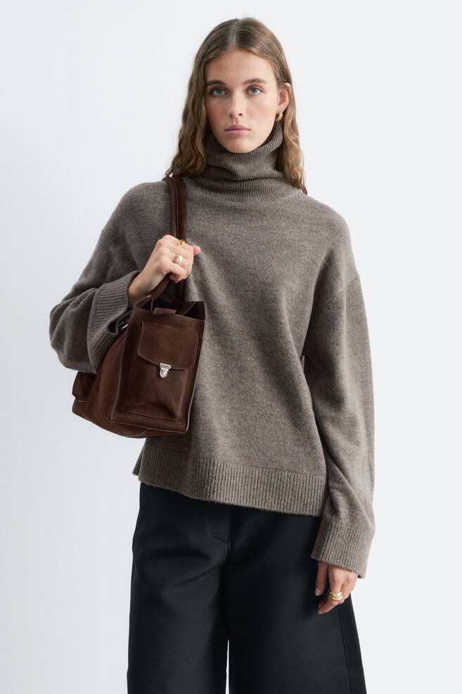Un pull col roulÃ© en mÃ©lange laine-coton, tricotÃ© dans un taupe chinÃ©, est portÃ© par le mannequin. Il prÃ©sente un col montant cÃ´telÃ©, des Ã©paules tombantes et des finitions cÃ´telÃ©es aux poignets et Ã  lâourlet. Le pull est associÃ© Ã  un pantalon noir, et le mannequin porte un sac bandouliÃ¨re en daim marron foncÃ© sur un bras. - 1