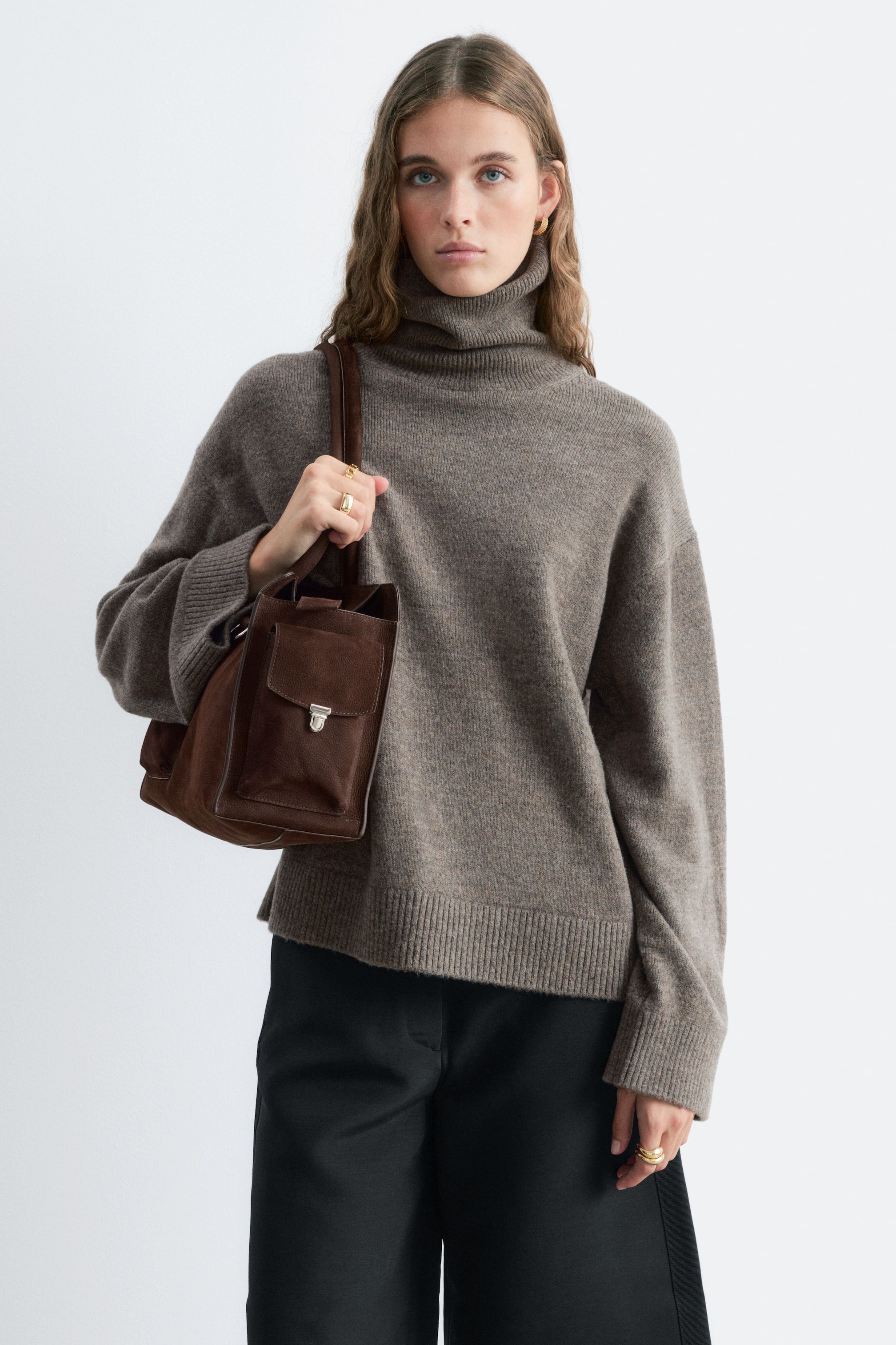 Größeres Bild anzeigen: Ein Rollkragenpullover aus Woll-Baumwoll-Melange in Taupe wird vom Model getragen und zeichnet sich durch einen hohen, gerippten Kragen, überschnittene Schultern sowie gerippte Abschlüsse an Ärmeln und Saum aus. Der Pullover wird zu einer schwarzen Hose kombiniert, und das Model trägt eine dunkelbraune Schultertasche aus Veloursleder über einem Arm.