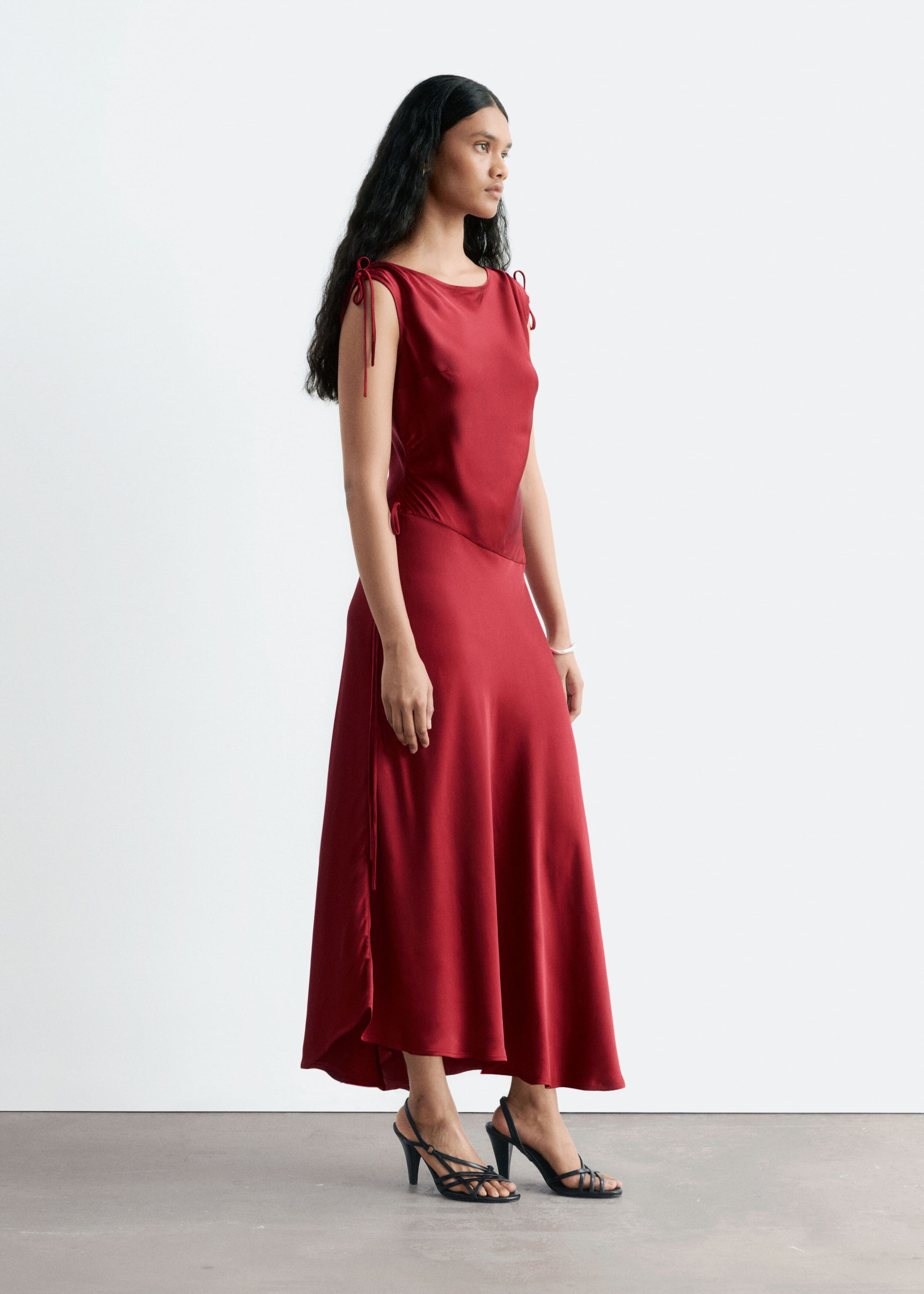 The model is wearing a red satin draped dress featuring drawstring ties at the shoulders and a gathered, asymmetric dropped waist. The sleeveless midi-length dress is paired with black strappy high-heeled sandals.