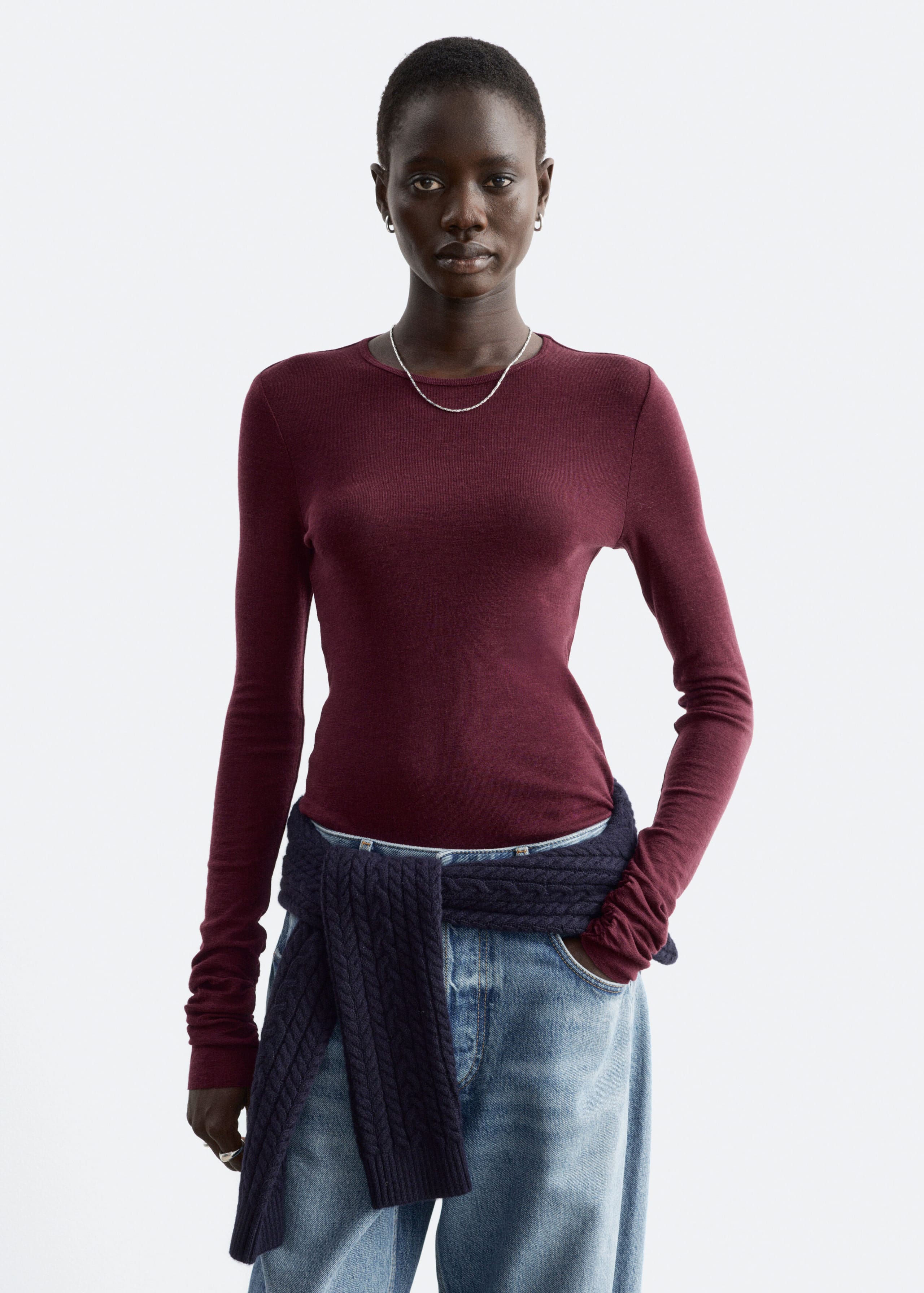 Ein bordeauxrotes, glatt gestricktes Wolloberteil mit Rundhalsausschnitt und langen Ärmeln wird vom Model getragen. Das schmal geschnittene Oberteil ist mit hellblauen Jeans und einem dunkel gefärbten, im Zopfmuster gestrickten Pullover kombiniert, der um die Taille gebunden ist.