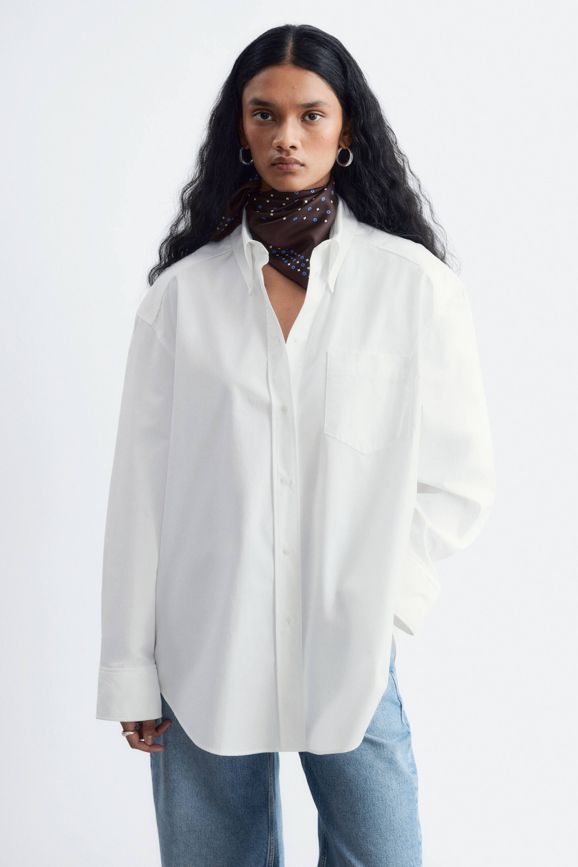 Visualizza l'immagine ingrandita: Una camicia bianca oversize in cotone, con abbottonatura, taschino sul petto e polsini trapuntati, indossata dalla modella. La camicia è portata fuori dai pantaloni sopra jeans azzurri ed è abbinata a una sciarpa da collo con fantasia marrone scuro.