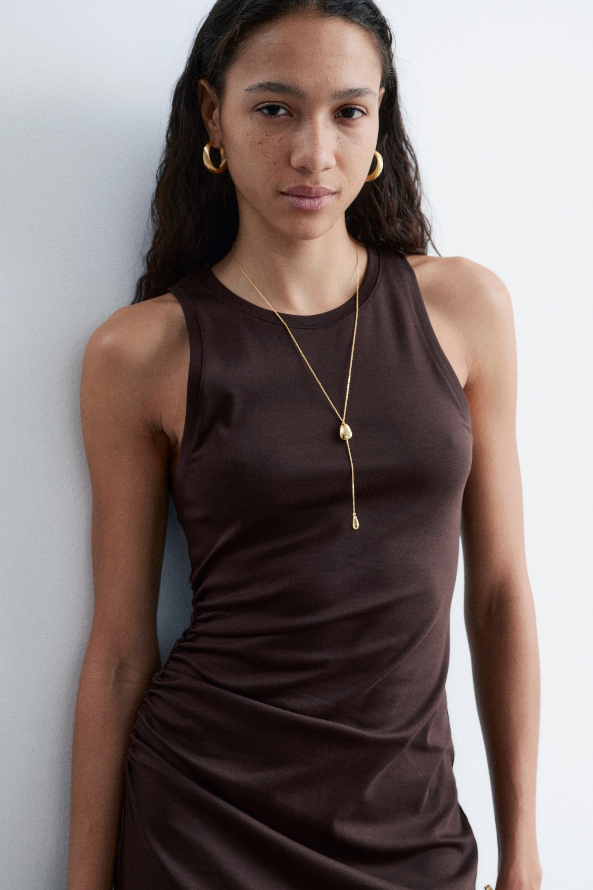 View larger image: A brown sleeveless dress featuring a round neckline and a fitted silhouette, with ruched detailing gathered on its left side.