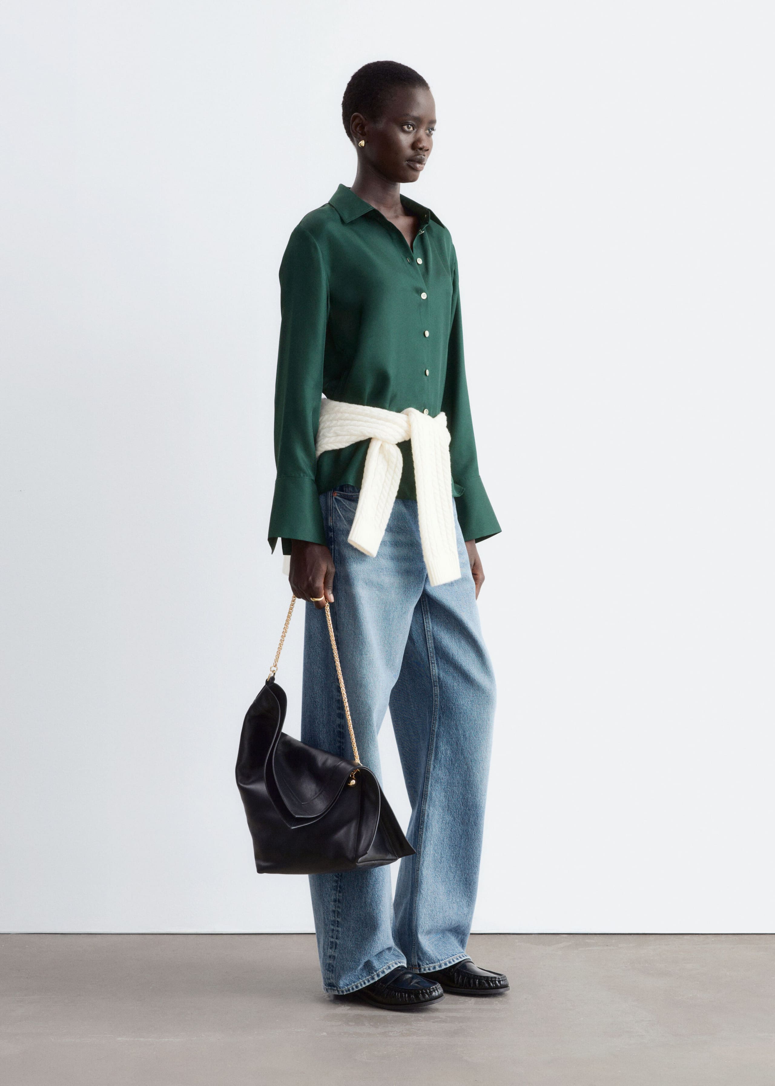 A dark green silk shirt featuring a classic collar, a button-down front, and wide cuffs is being worn by the model. The shirt is styled with a white knit jumper tied around the waist, wide-leg blue jeans, and a black leather handbag being carried by the model.
