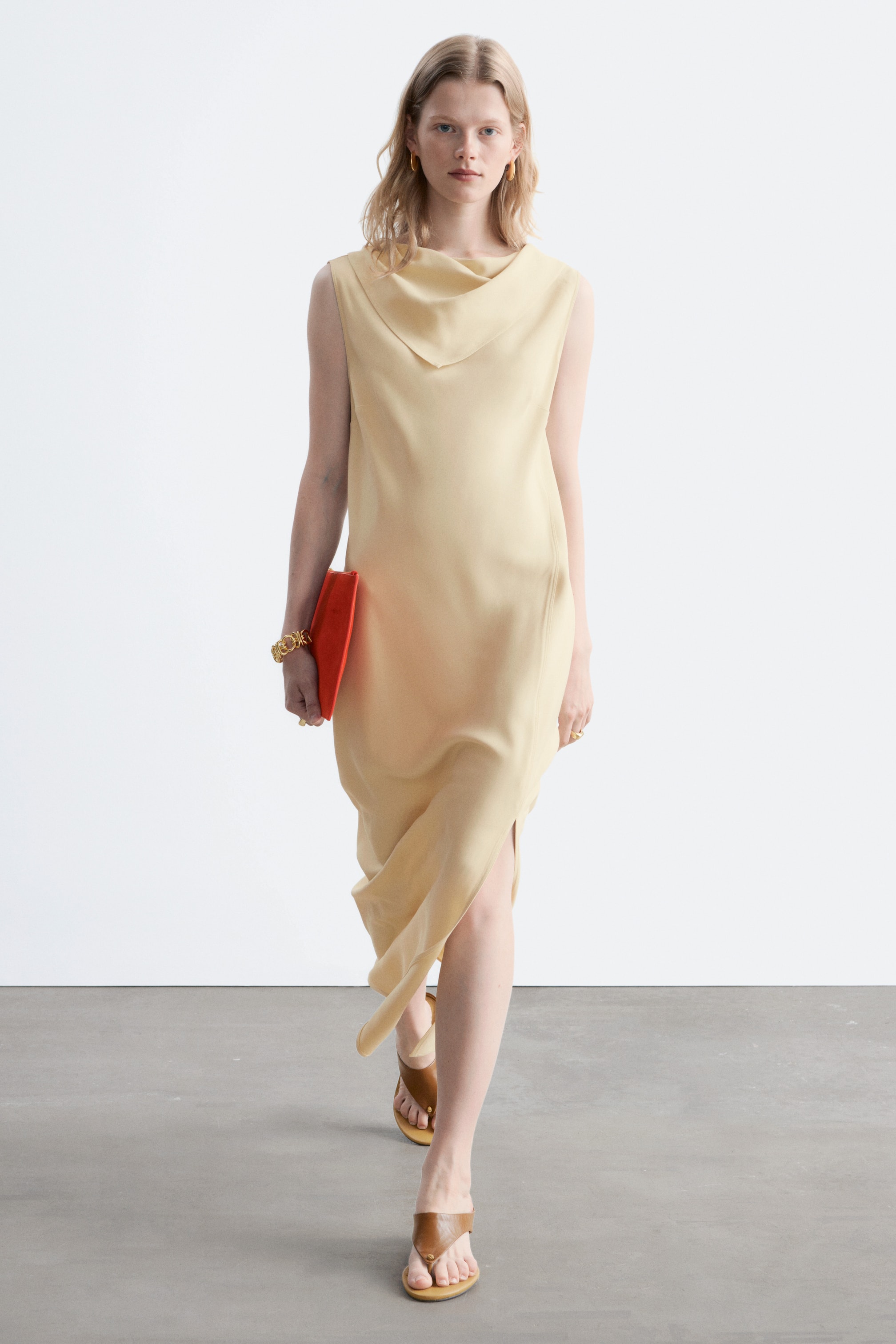 Ver imagen más grande: Un vestido midi sin mangas de color amarillo pálido presenta un escote drapeado y una abertura lateral, fluyendo con elegancia al moverse hacia adelante.