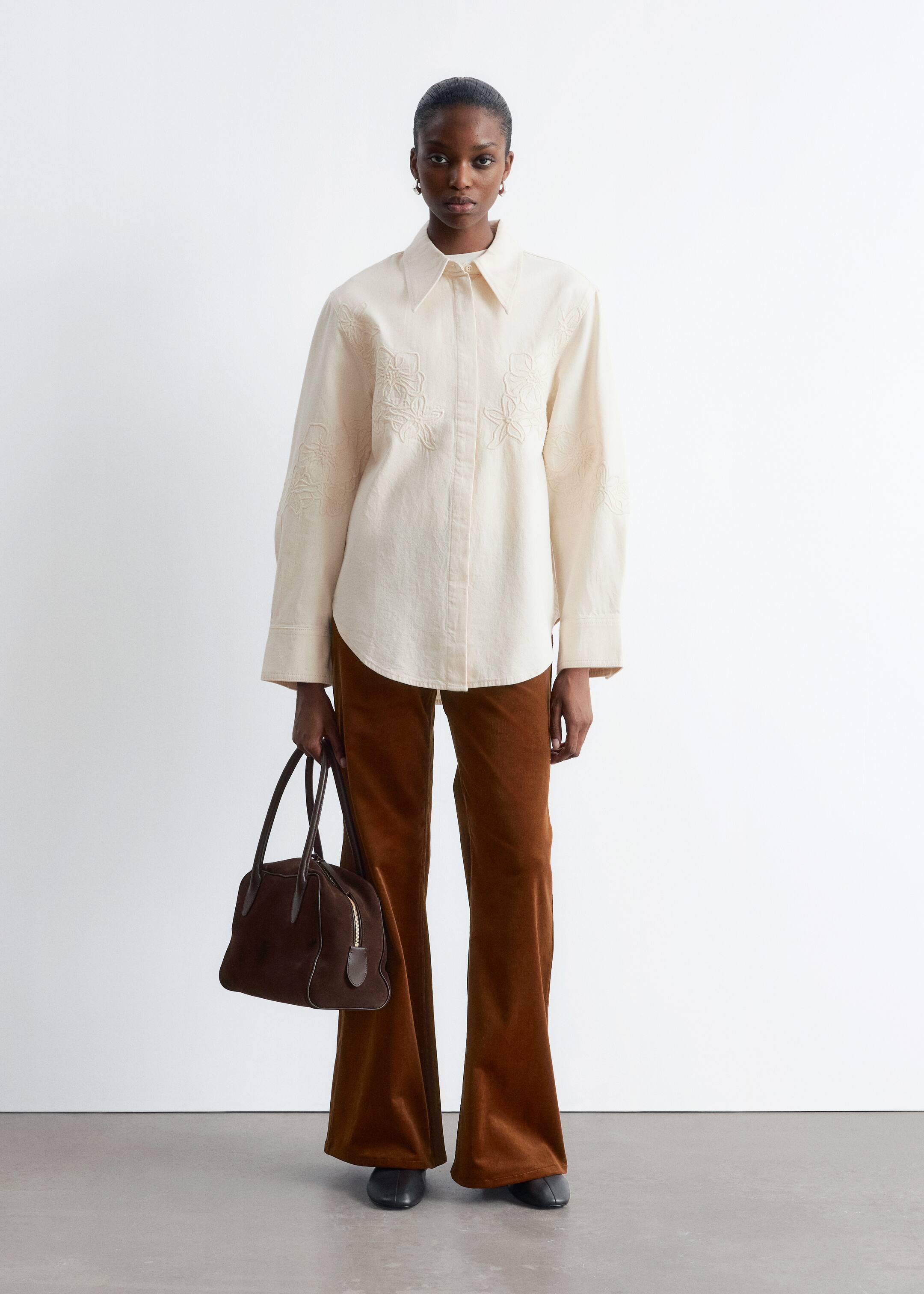 Zobrazit větší obrázek: Modelka má na sobě krémově bílou, volně padnoucí džínovou košili, která je zdobena vyšívanými květinovými ozdobami na hrudi a rukávech, a má klasický límec.