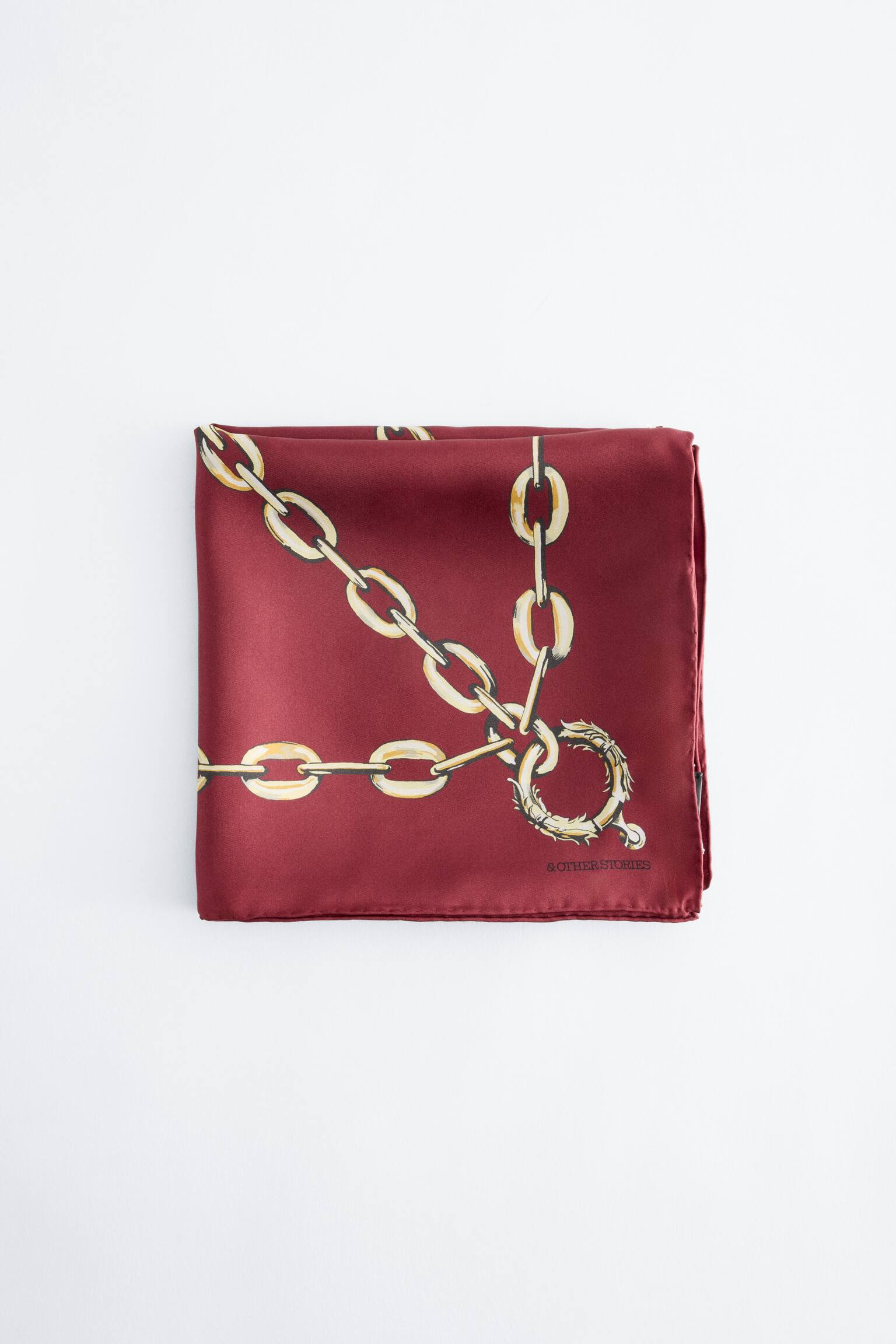 Un foulard quadrato in seta color bordeaux Ã¨ appoggiato piegato e disteso, con una stampa grafica di grandi maglie di catena dorate e unâornata chiusura circolare, con il nome del brand â& OTHER STORIESâ visibile nellâangolo in basso a destra.