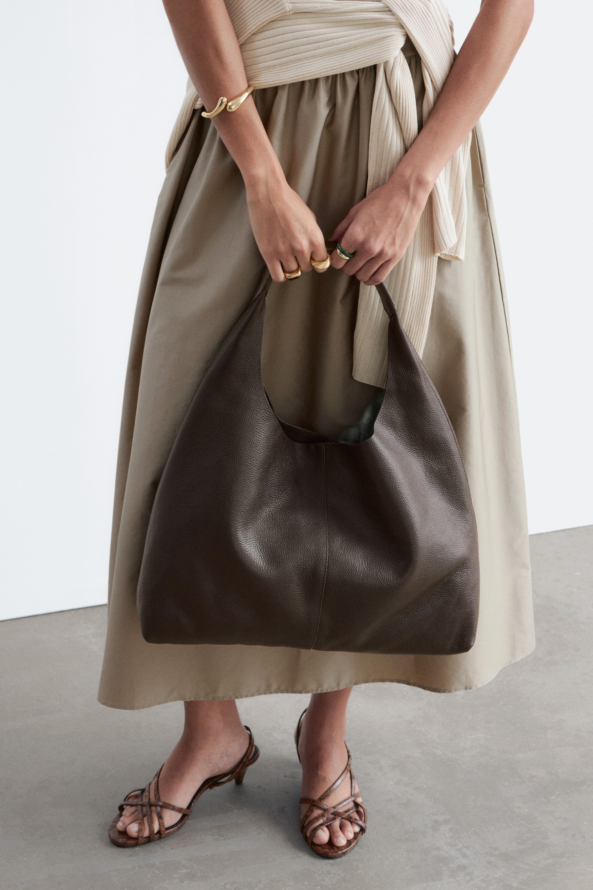 Visualizza immagine più grande: Una grande borsa tote in pelle marrone scuro con una morbida grana è tenuta dalla modella, che afferra il suo unico manico.