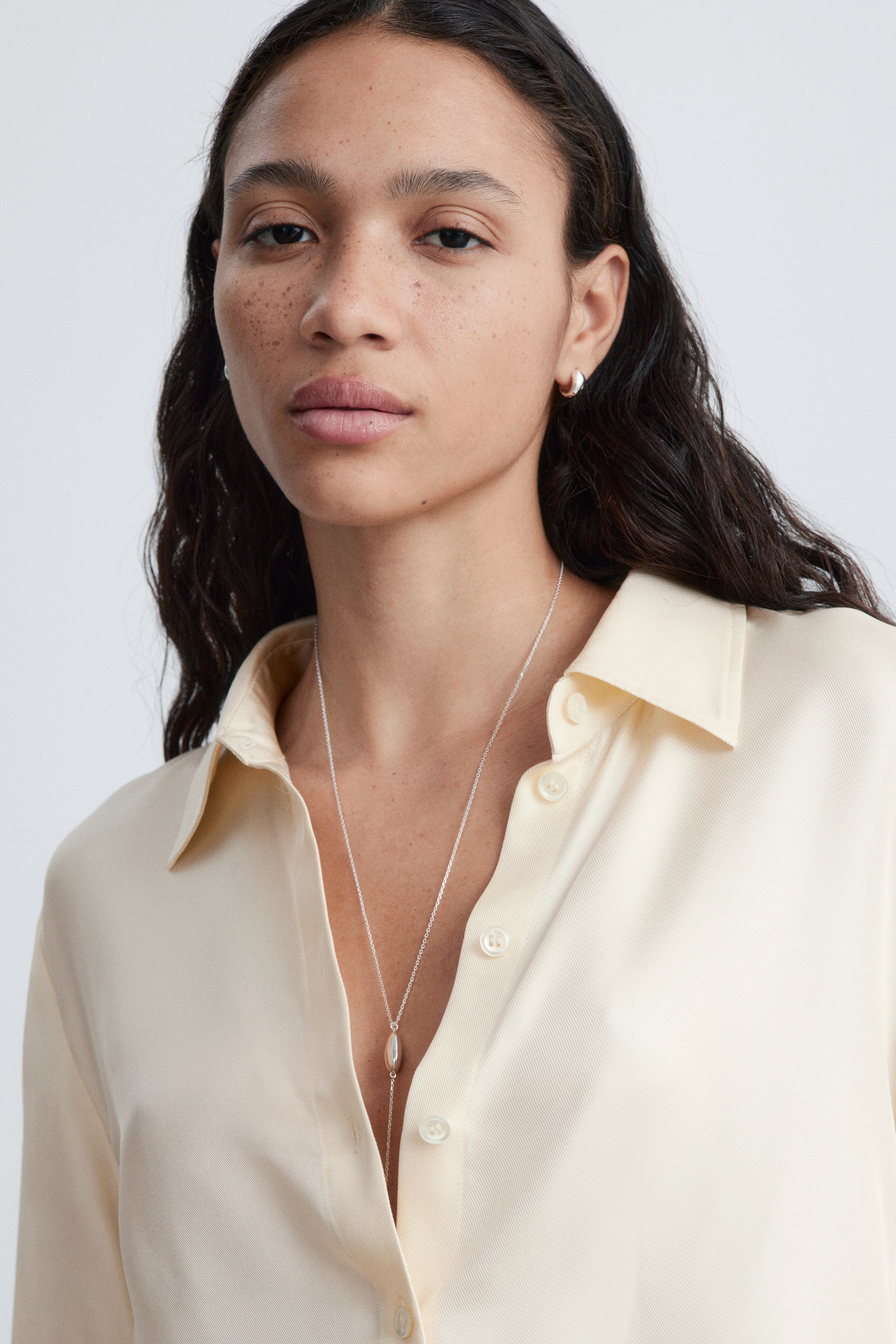 Agrandir l'image: De petites boucles d'oreilles créoles argentées sont portées sur les lobes des oreilles. Un long collier à chaîne argentée avec un pendentif allongé et lisse est drapé sur une chemise crème à col ouvert.