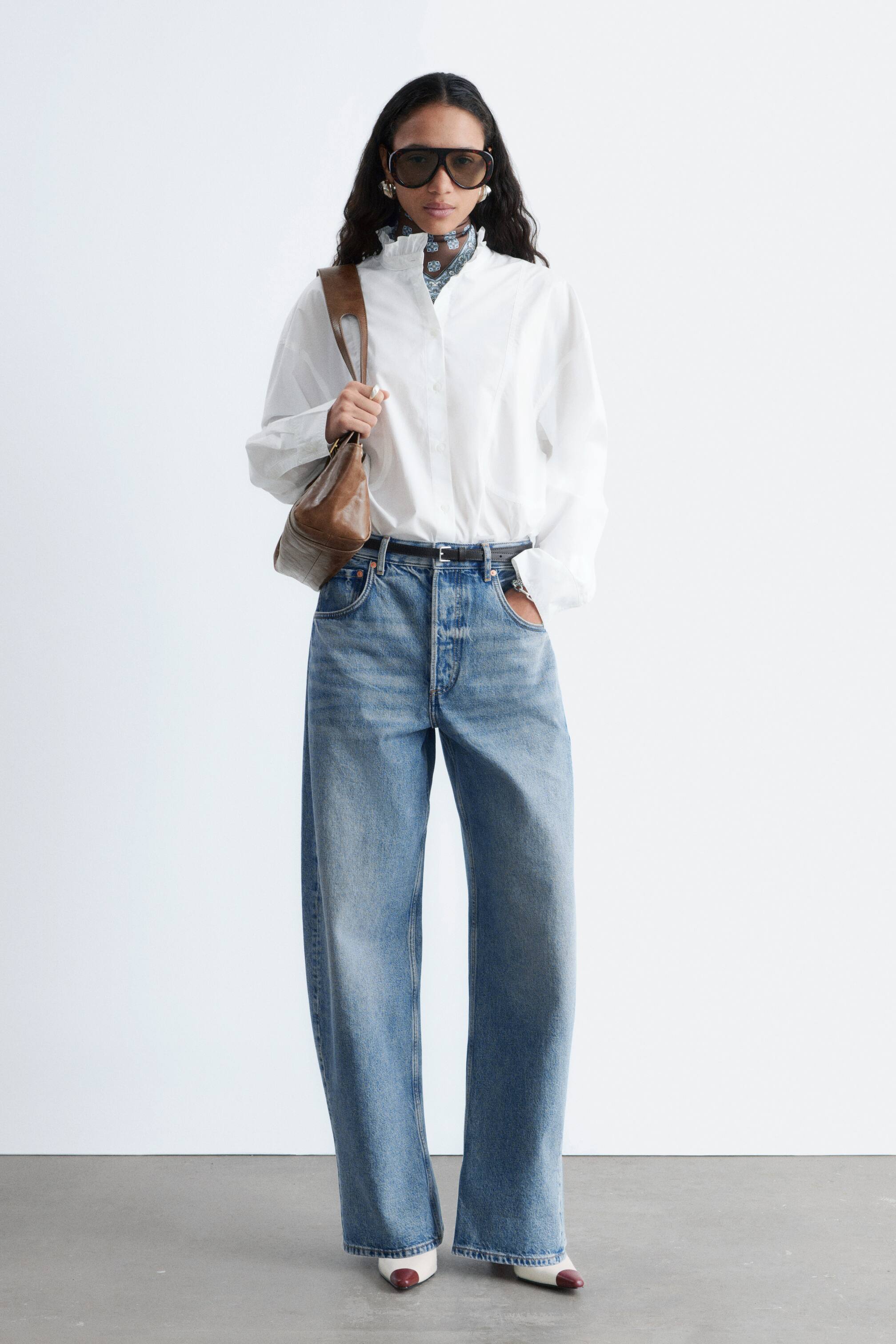 Ingrandisci l'immagine: I jeans lavaggio azzurro chiaro presentano un design a vita alta con una chiusura a bottoni visibile, un taglio ampio e largo che si estende fino al pavimento, e tasche standard anteriori e posteriori con una cintura nera che attraversa i passanti.