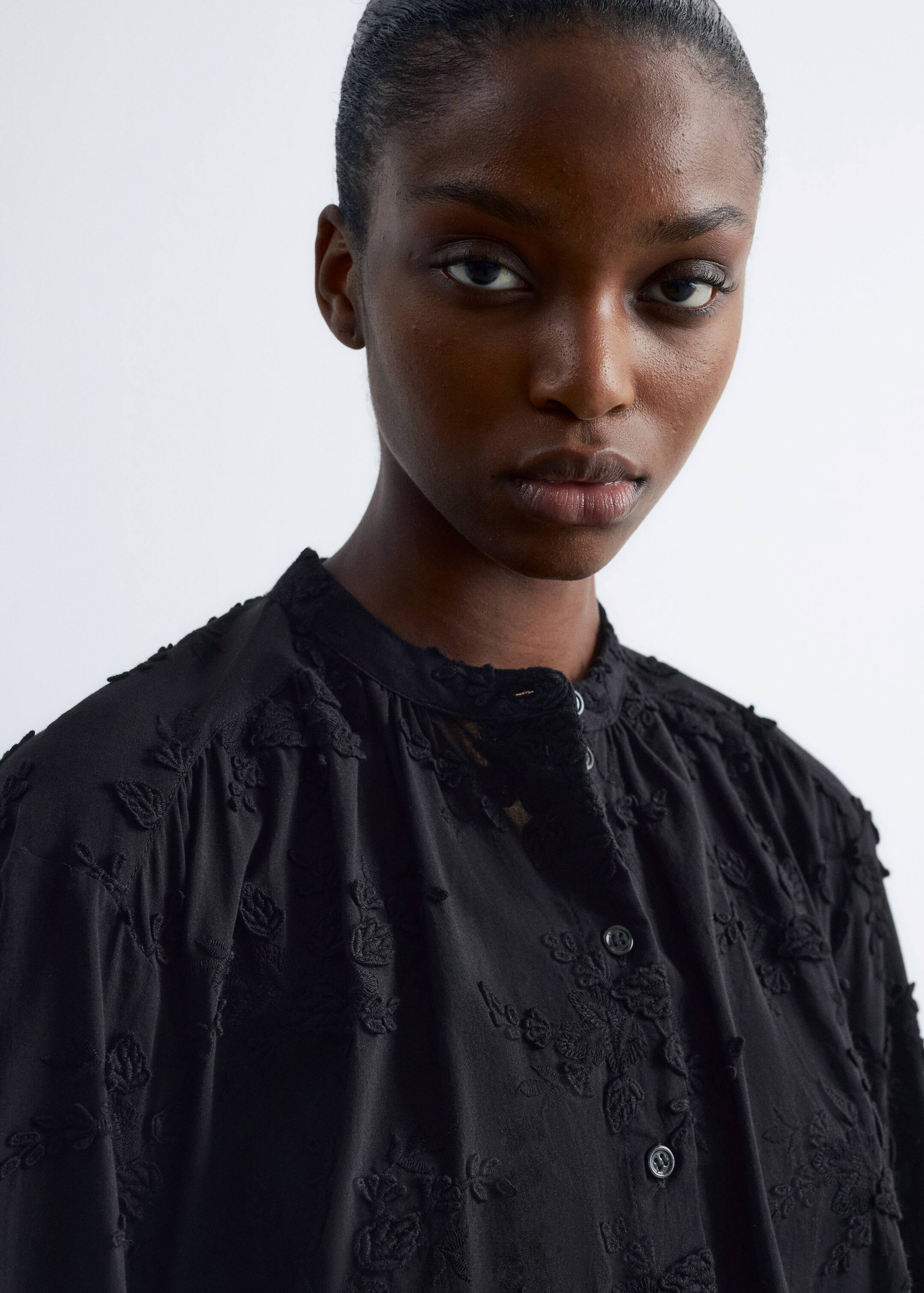 Ver imagem maior: Uma blusa preta apresenta uma gola mandarim e uma carcela de botões. É adornada com bordados em relevo de flores e folhas pretas tonais, e o tecido está franzido nos ombros, indicando um ajuste descontraído.