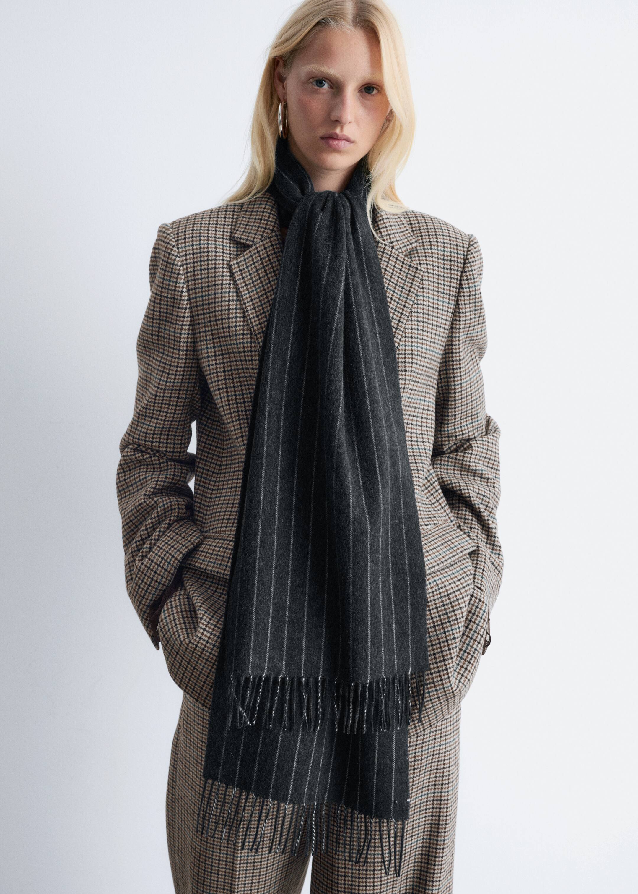 Größeres Bild anzeigen: Ein dunkelgrauer Nadelstreifenschal ist um den Hals gebunden und hängt vertikal über einem karierten Blazer. Er besitzt eine gebürstete Textur mit dünnen weißen Nadelstreifen und ausgefransten Enden.