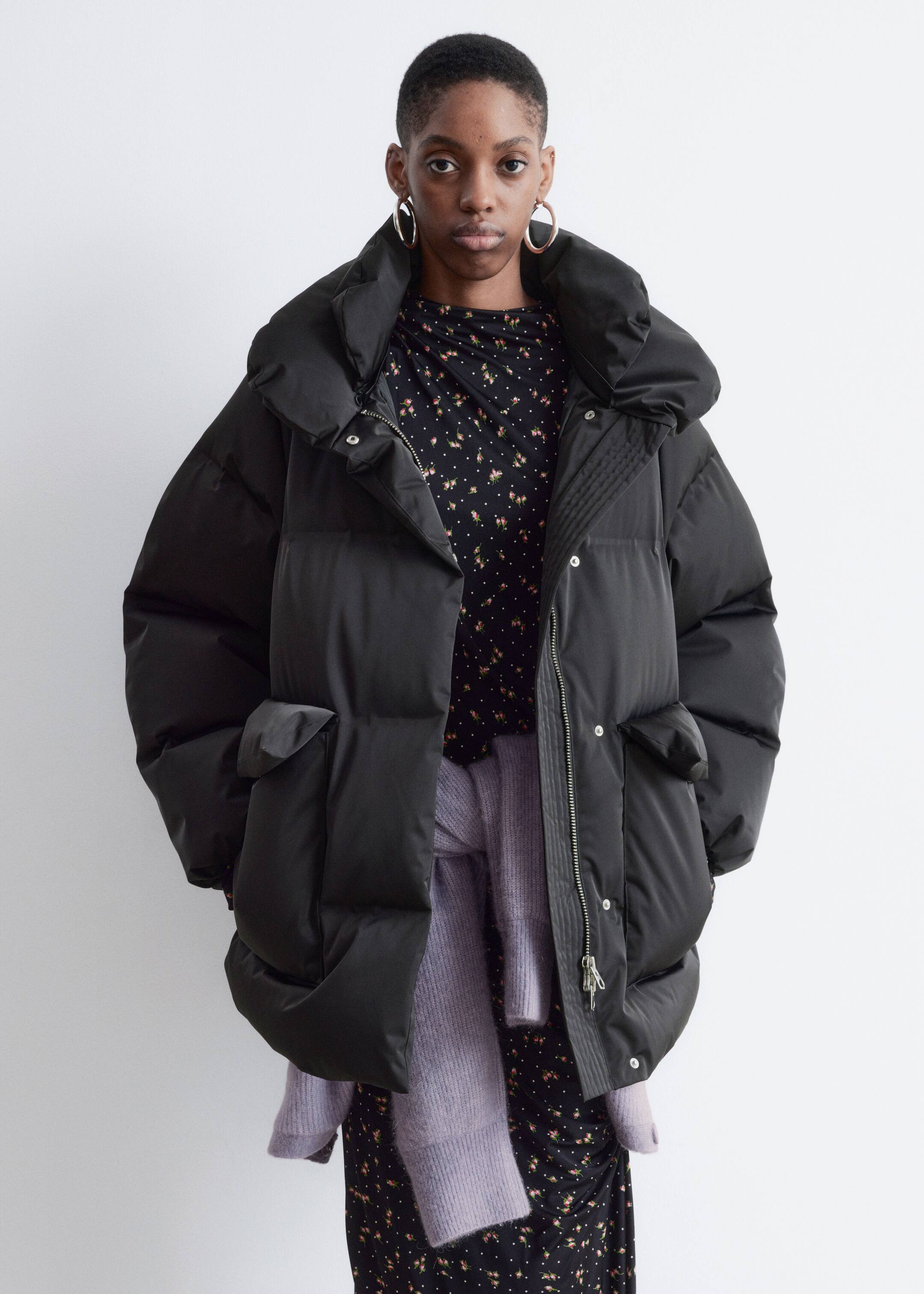 View larger image: A black puffer jacket is presenting front-facing, featuring an oversized fit, a high padded collar, and a full-zip front closure with snap buttons. It is displaying two flap pockets, with a floral dress visible underneath the open jacket and a lavender sweater tied around the waist.