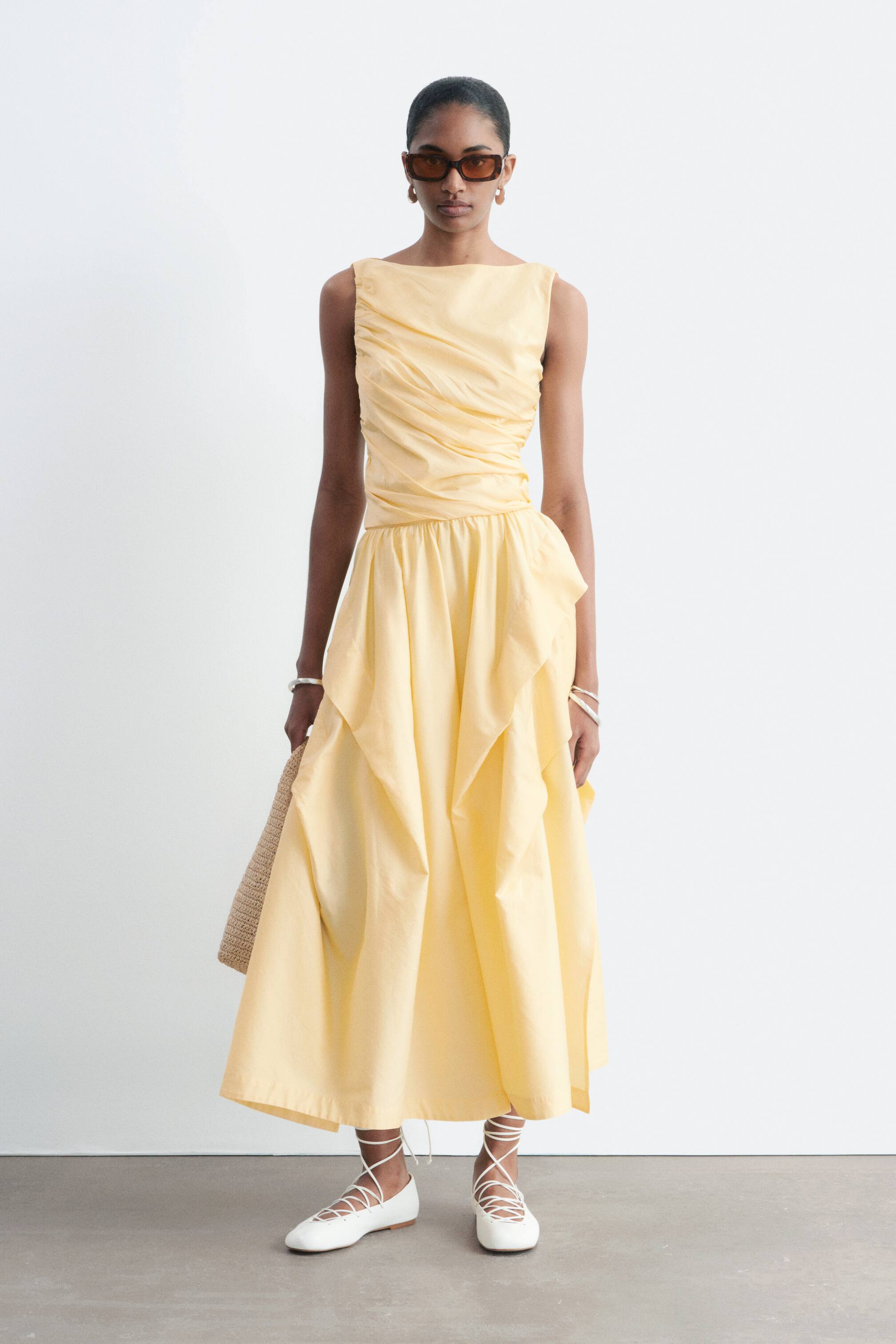 Zobrazit větší obrázek: Žluté midi šaty představují nabíraný, bezrukávový živůtek se širokým výstřihem. Jejich splývavá sukně je navržena s vrstvenými, asymetrickými látkovými panely pod staženým pasem.