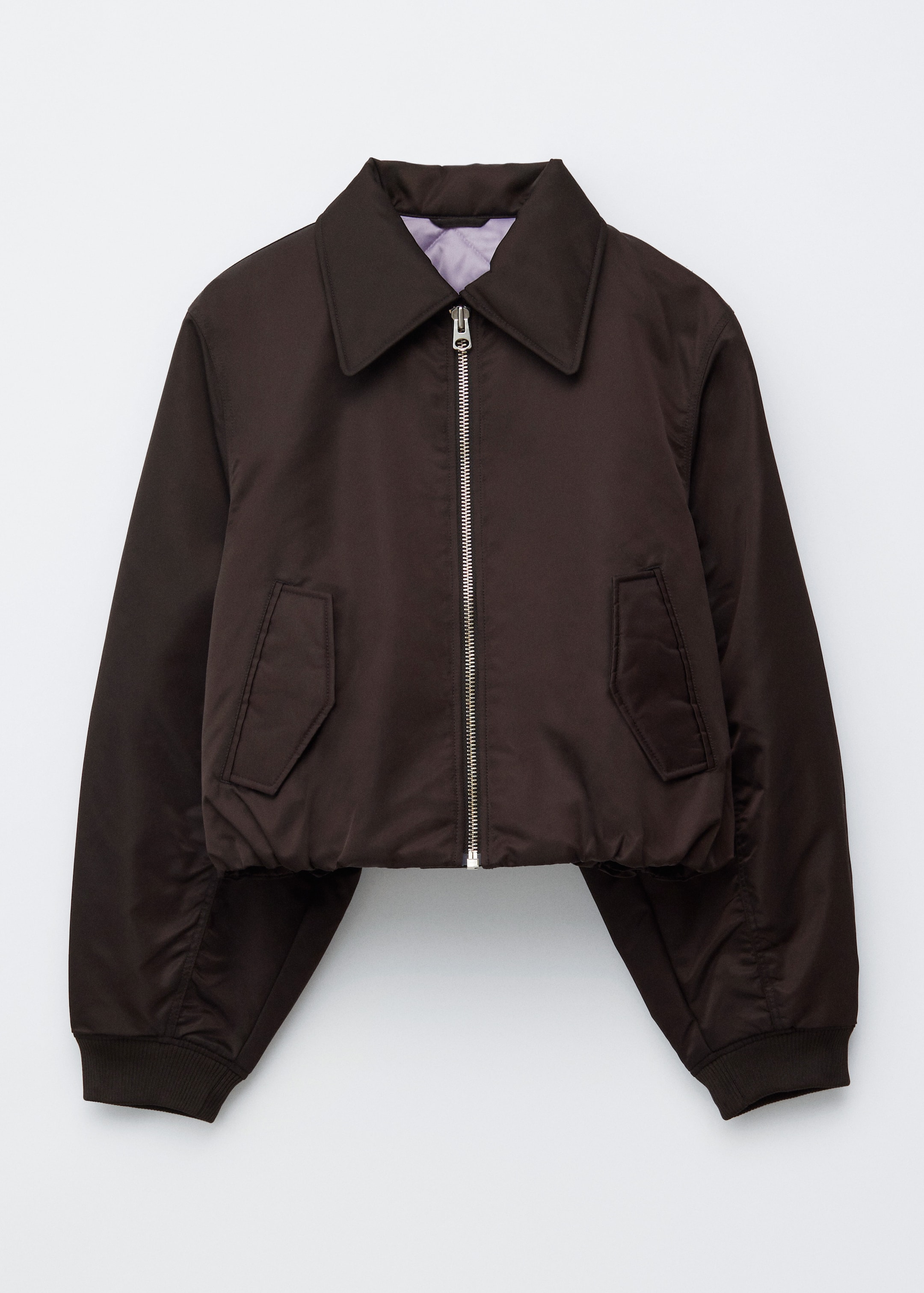 Ingrandisci l'immagine: Una giacca bomber marrone scuro viene mostrata dal davanti, caratterizzata da una silhouette squadrata e corta con un colletto classico, una chiusura con zip color argento e polsini a coste. Due tasche con patta inclinate sono visibili sul davanti, e una fodera trapuntata color viola chiaro è visibile al colletto.