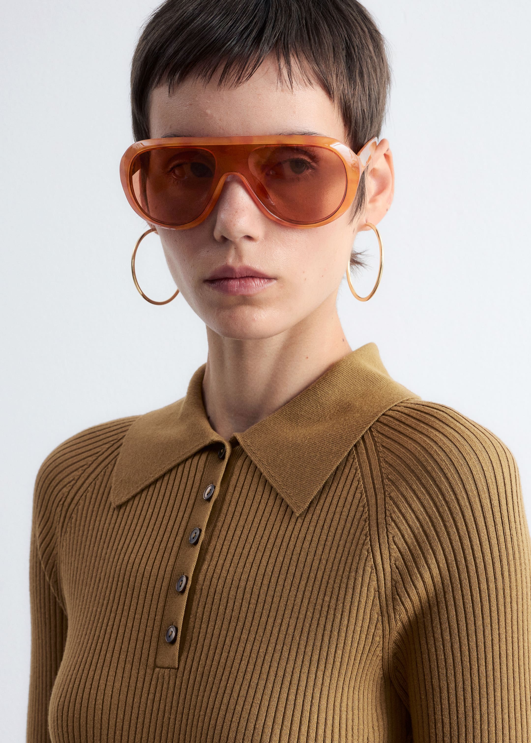 Agrandir l'image: Des lunettes de soleil surdimensionnées de style aviateur en ambre translucide avec des verres teintés sont portées, avec un double pont distinct. Un polo en maille côtelée de couleur camel est également porté, avec un col pointu, une patte de boutonnage avec cinq boutons visibles et des manches raglan.