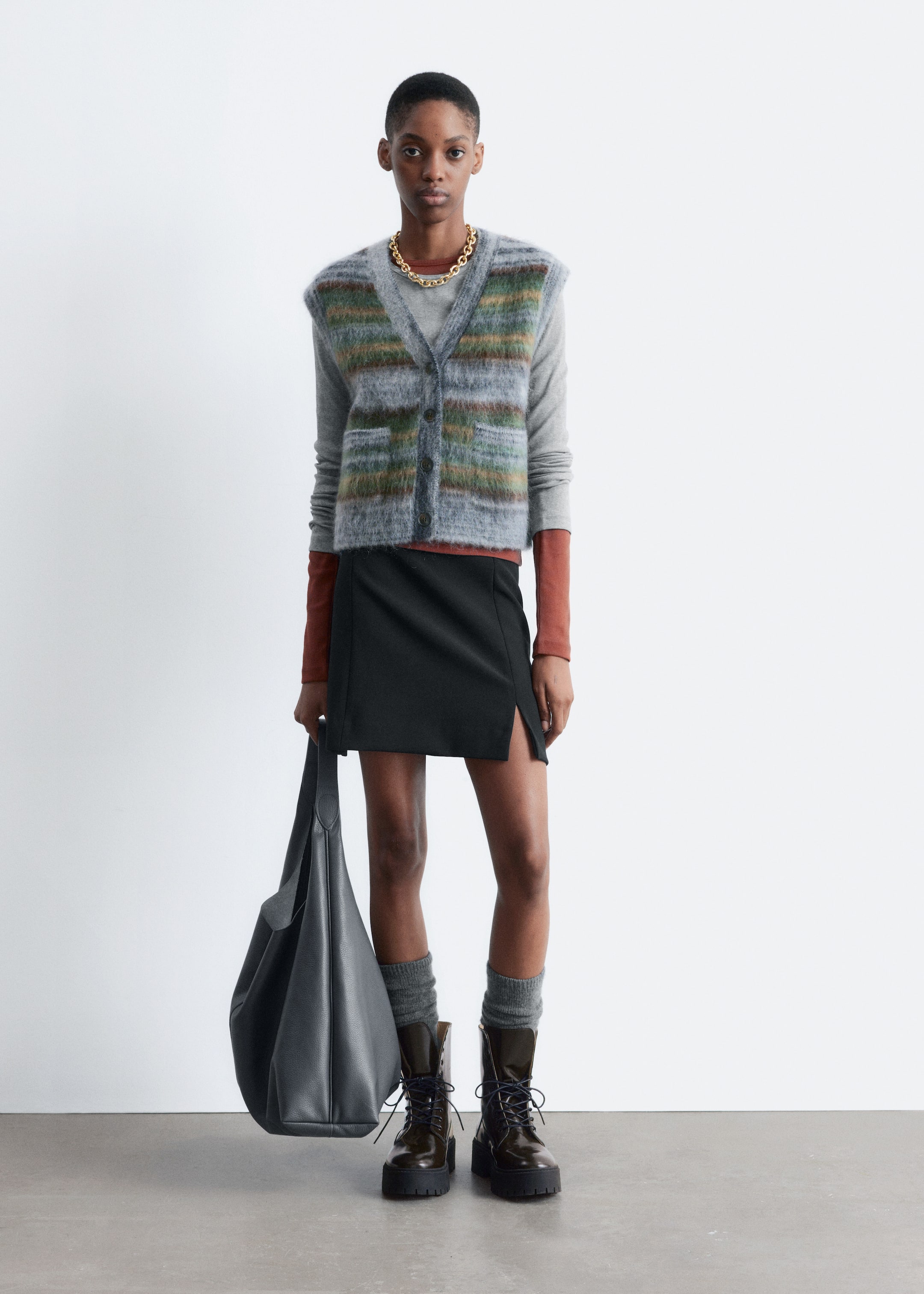 Visualizza immagine più grande: Un gilet in misto mohair e lana, caratterizzato da una finitura spazzolata e tattile e da righe orizzontali in tonalità di grigio, verde, marrone e ruggine, è indossato dalla modella. Ha uno scollo a V, bordi a coste, due tasche applicate frontali e una chiusura con bottoni sul davanti ed è sovrapposto a un top a maniche lunghe grigio chiaro e a un top a maniche lunghe color ruggine.