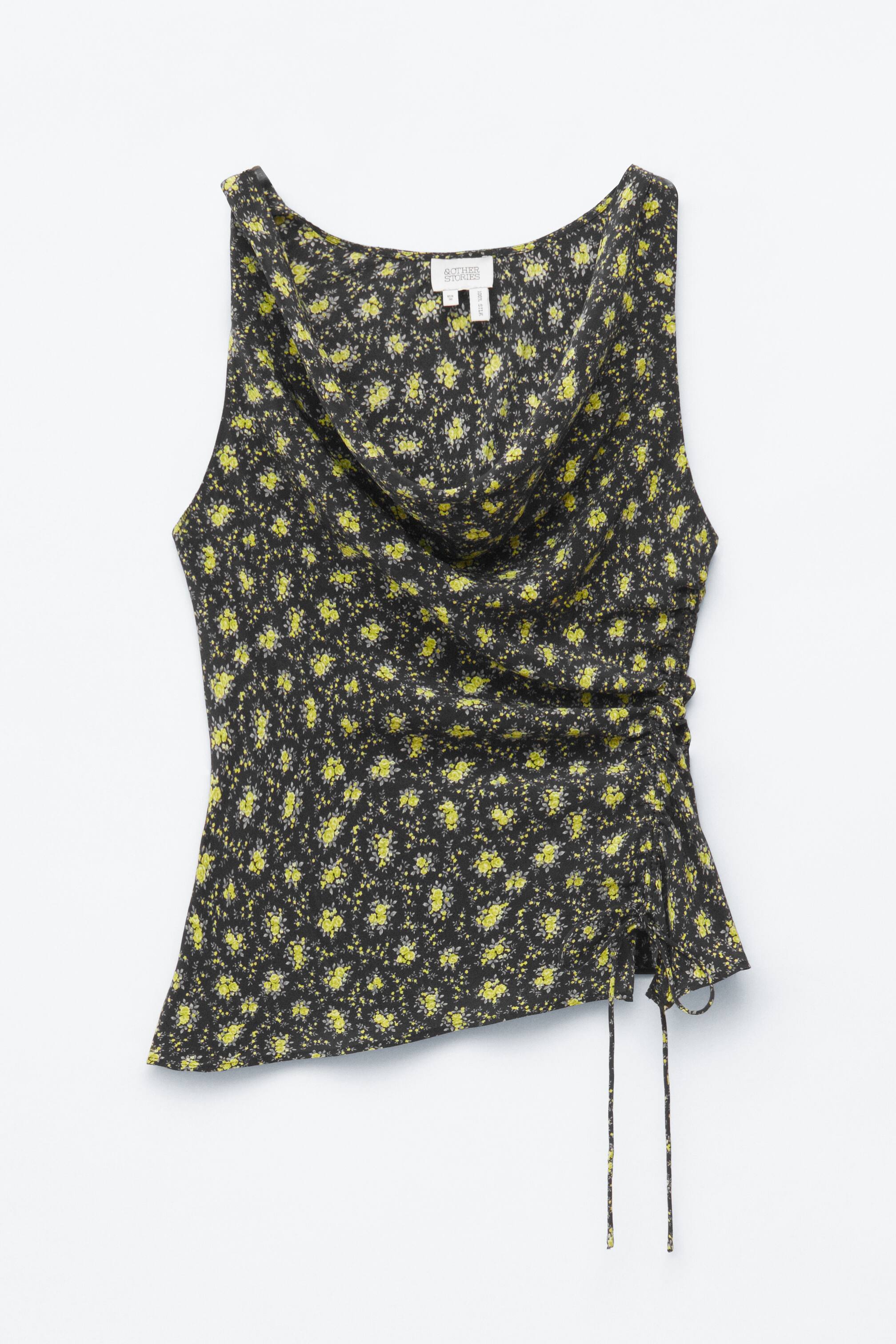 Visa större bild: En ärmlös topp med en mörk bakgrund och utspridda gula och ljusgrå blommönster. Den är designad med en kowlkrage och har rynkor med dragsnören på plaggens högra sida, vilket skapar en asymmetrisk nederkant.