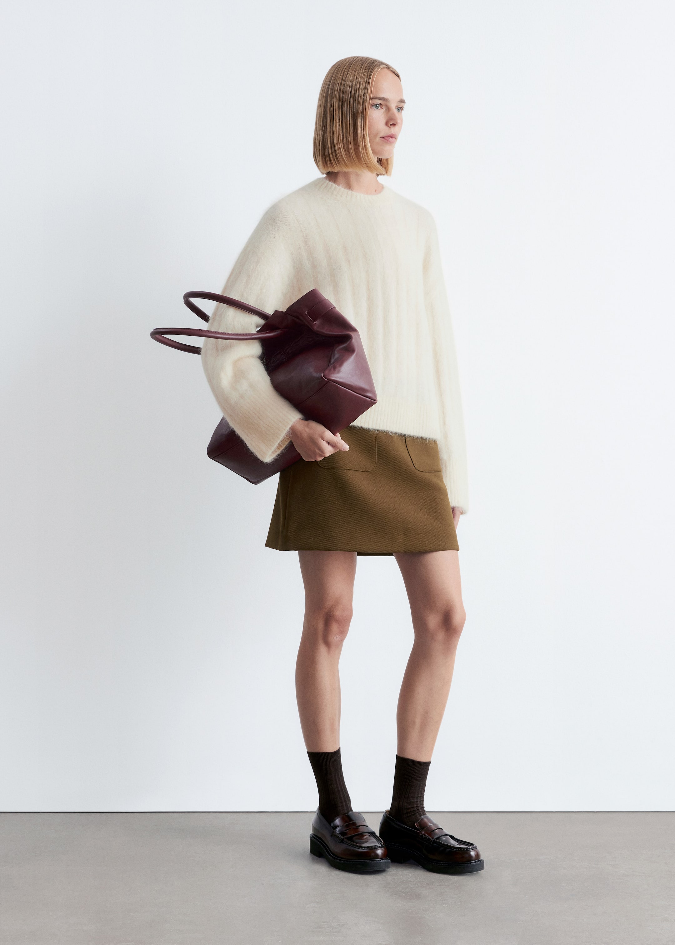 View larger image: A cream long-sleeved jumper, featuring a loose fit, fuzzy texture, subtle rib-knit, dropped shoulders, and ribbed crew neck, cuffs, and hem, is being worn by the model. The model is styling the jumper with an olive mini skirt and is holding a burgundy tote bag against its side.